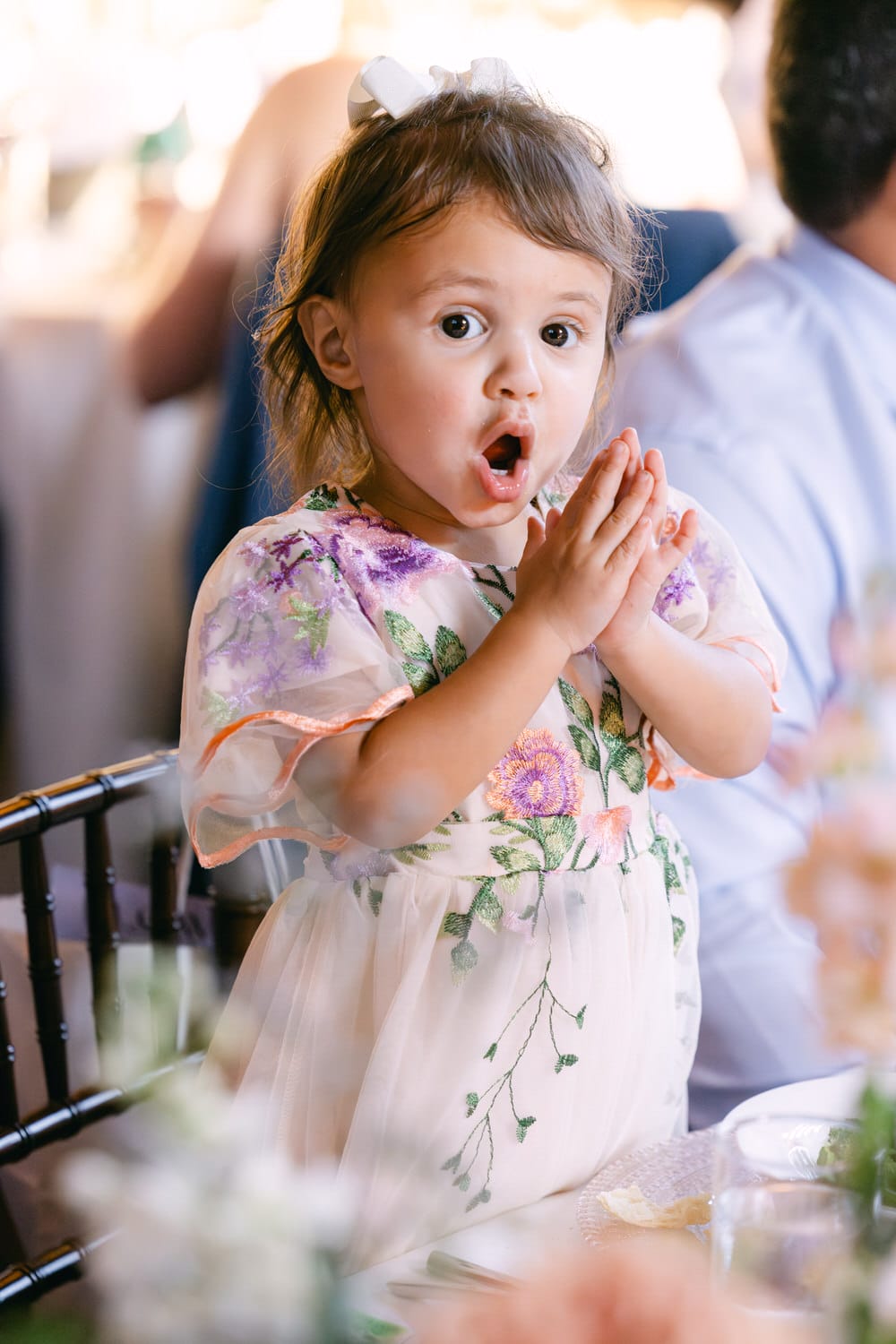 A young girl with a surprised expression, wearing a floral dress and a white bow in her hair, clapping her hands at a social gathering.