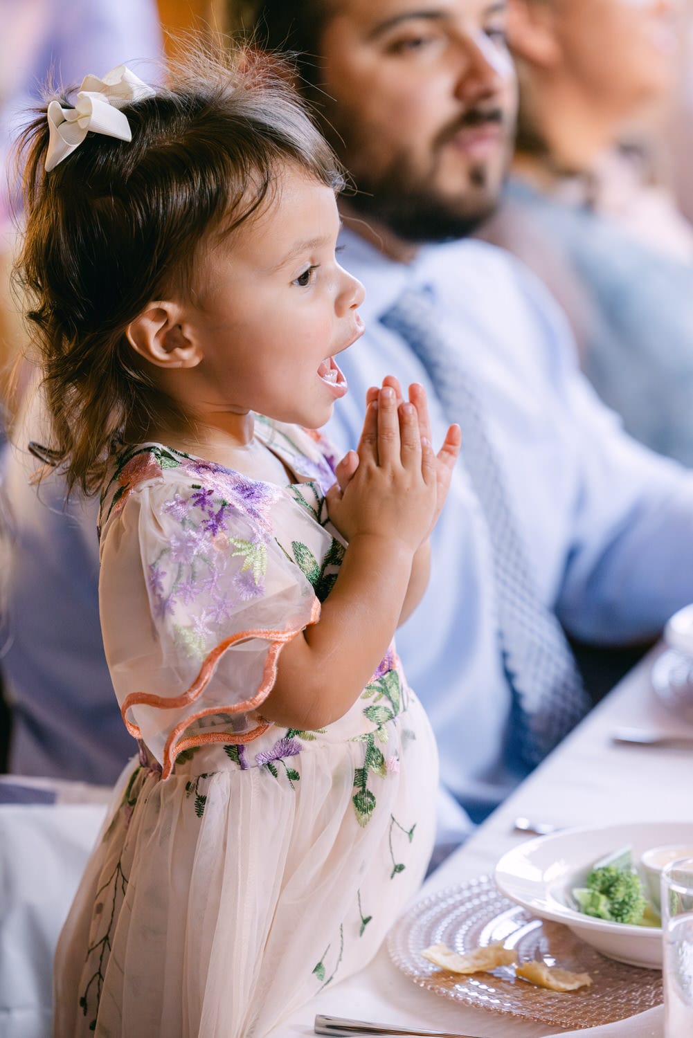 A young girl in a decorated dress with hands clasped in excitement at a dining table, with a partially visible man in the background.