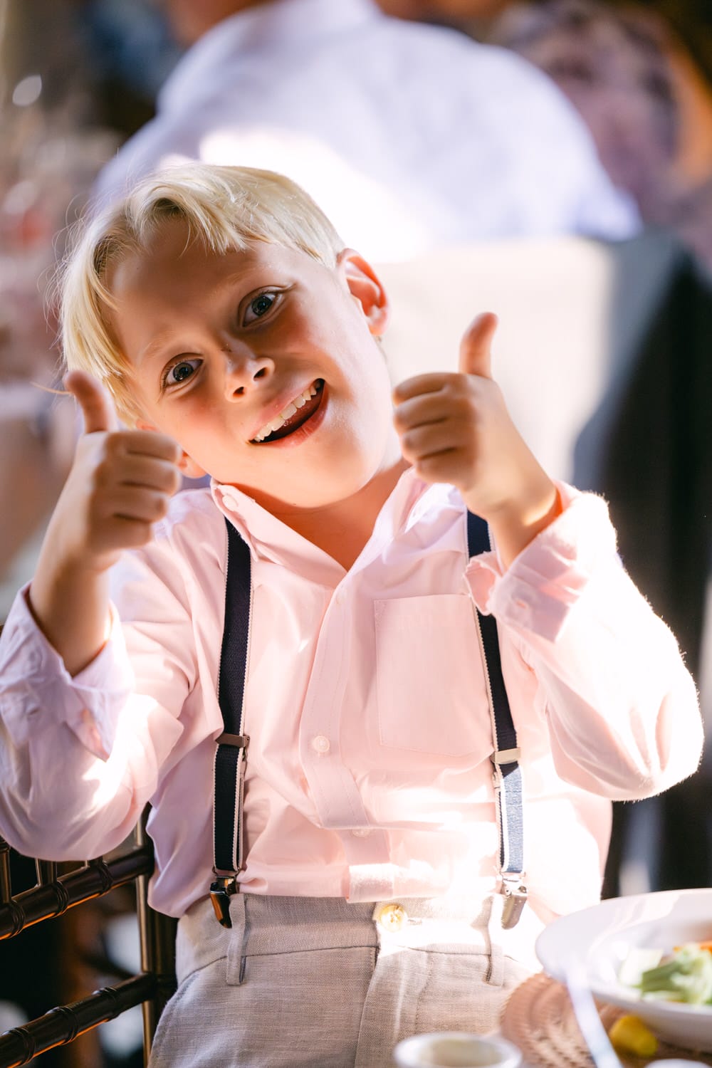 A smiling young boy in a pink shirt and suspenders giving two thumbs up at a dining event.