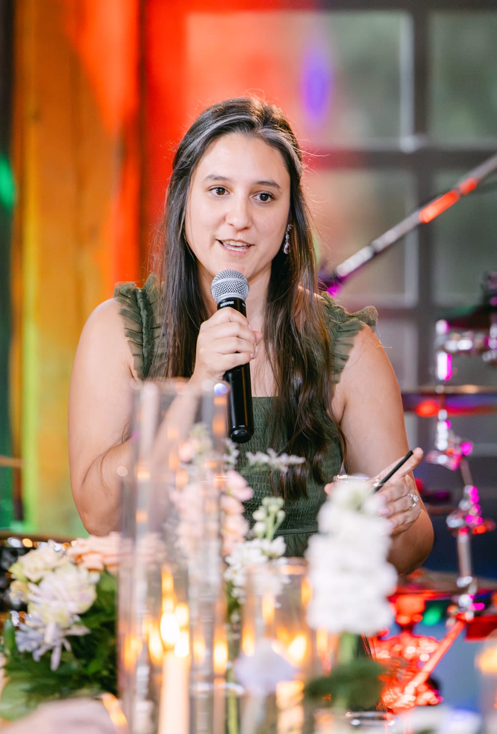 A woman speaking into a microphone with flowers and decorative lights in the foreground.