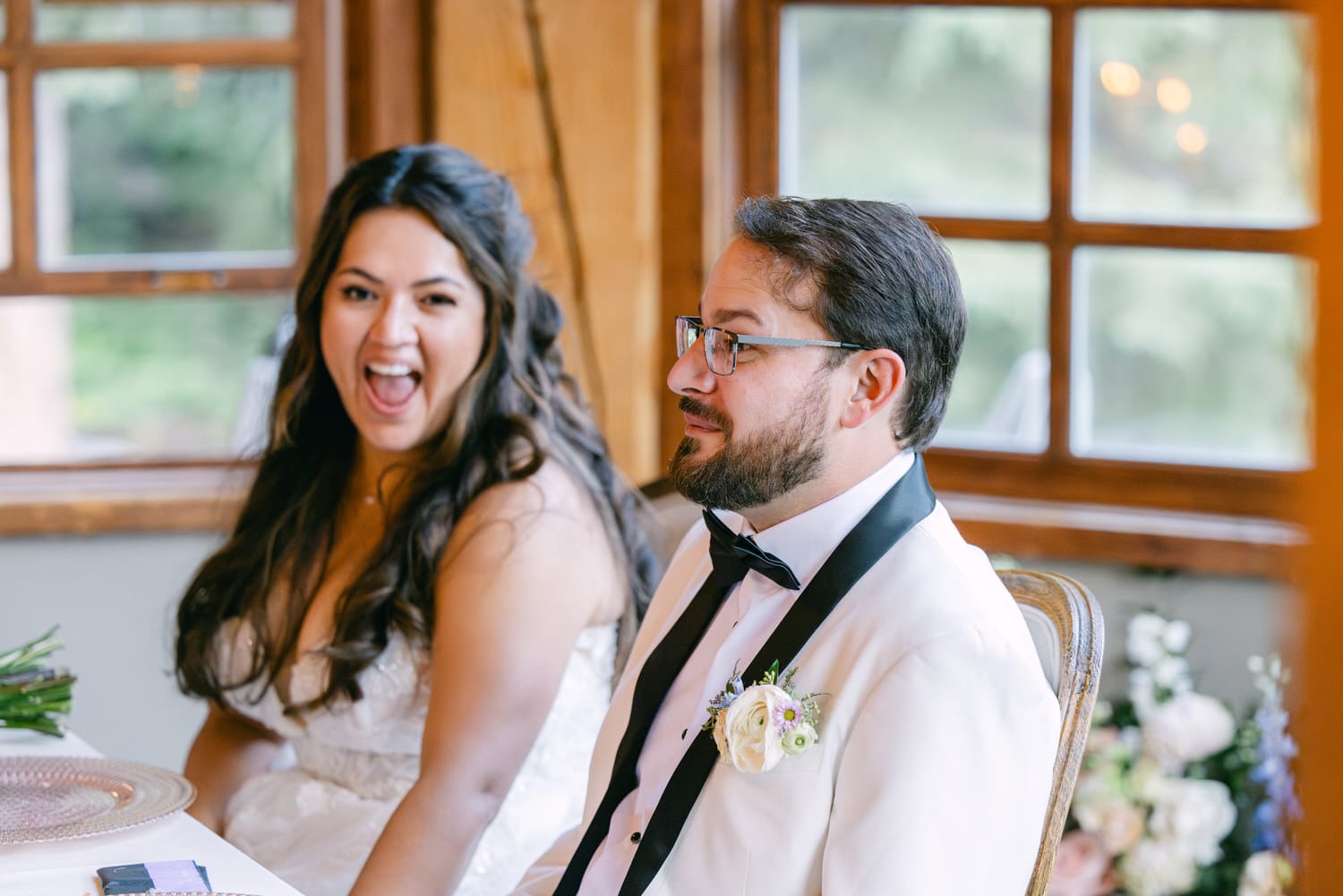 Bride laughing joyfully while sitting beside groom in a wedding reception setting with elegant decor and window in the background.