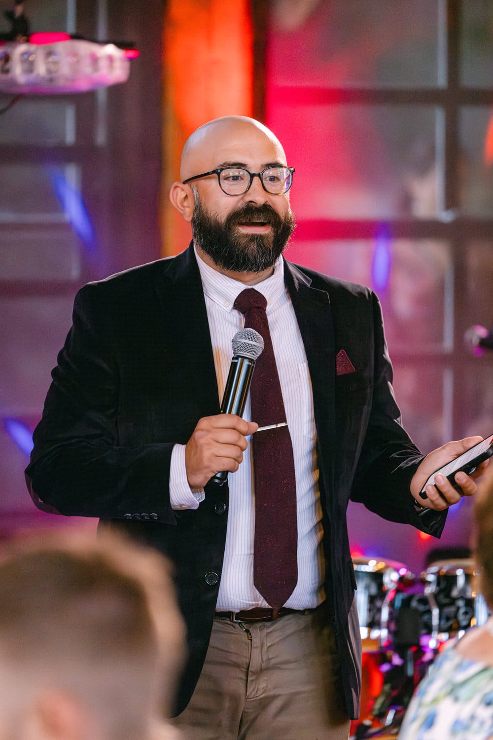 Man with a microphone speaking at an event, wearing glasses, a dark suit, and a red tie.