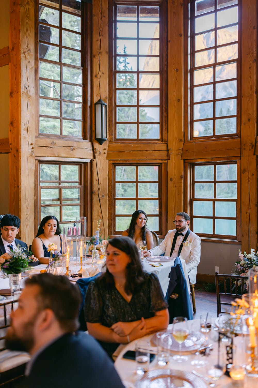 Guests seated at elegantly set tables inside a rustic venue with large wooden framed windows showcasing mountain views.