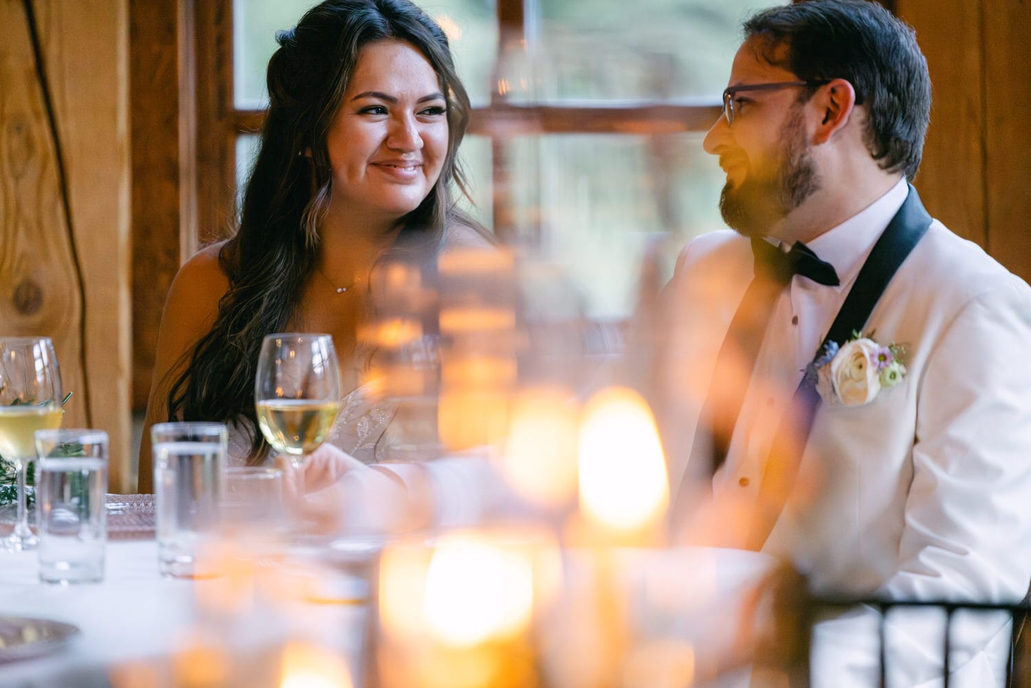 A bride and groom sharing a warm smile at a candlelit dinner table, with focus on the bride and soft bokeh effect from the candles in the foreground.