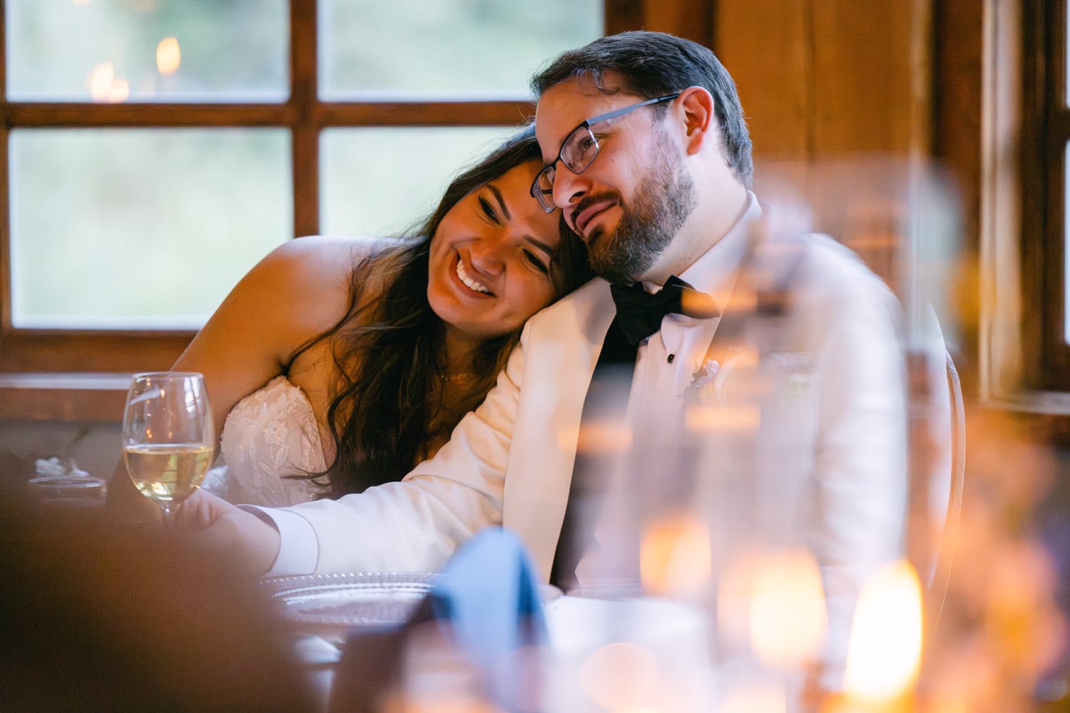 A couple in wedding attire lovingly leans on each other, smiling and enjoying a moment together, with soft lighting and a blurred foreground adding a cozy atmosphere.