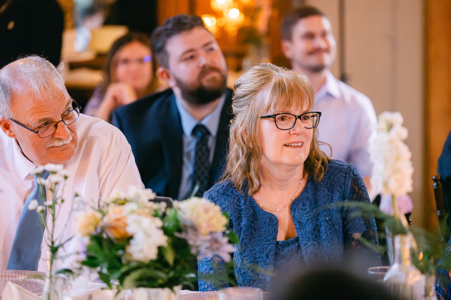 A group of guests at an event with focus on a smiling woman in blue, and a man in white looking towards a point of interest out of frame, with others in the background and floral arrangements on the table.