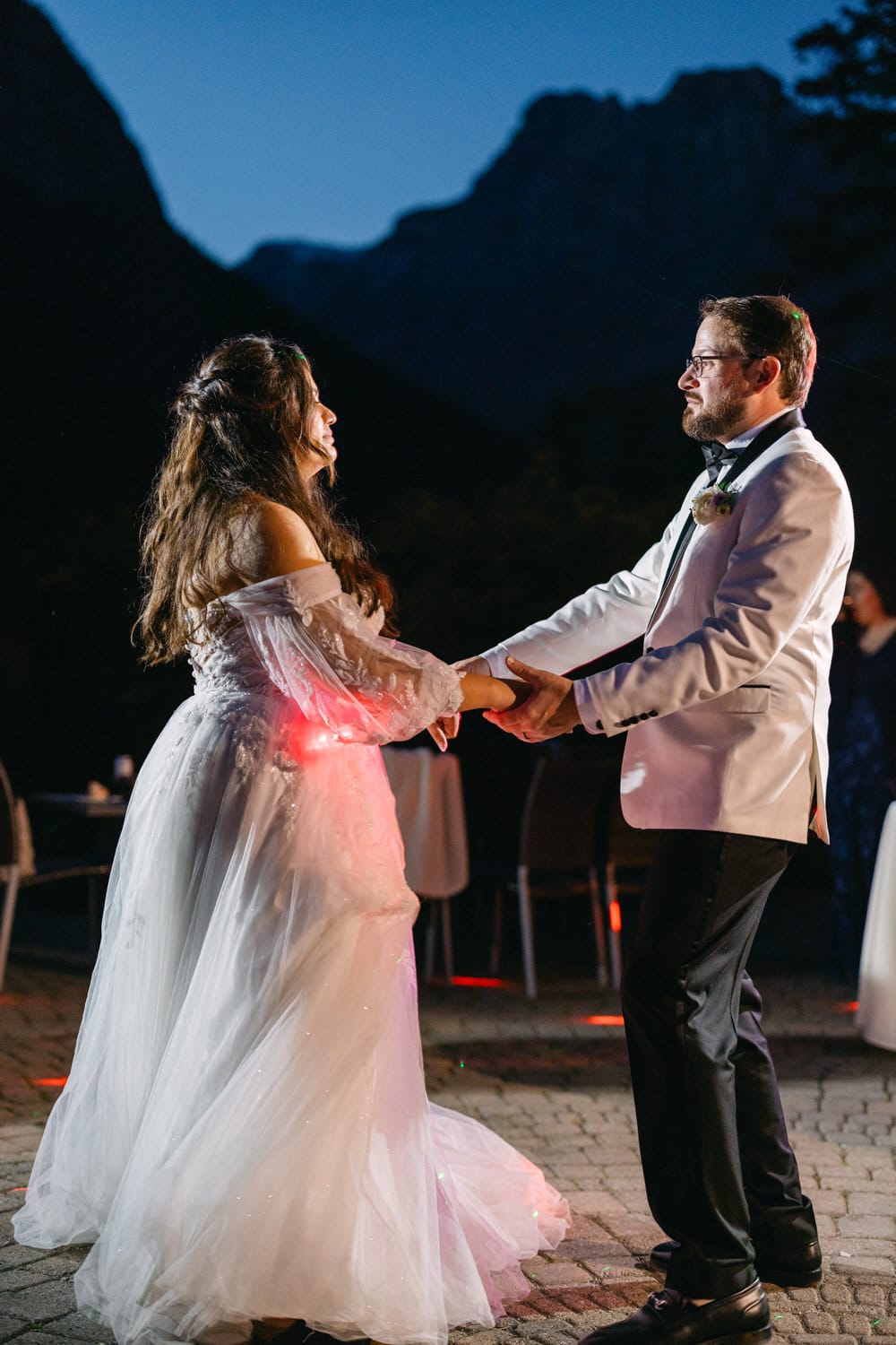 Bride and groom holding hands and dancing at night with dark mountains in the background.