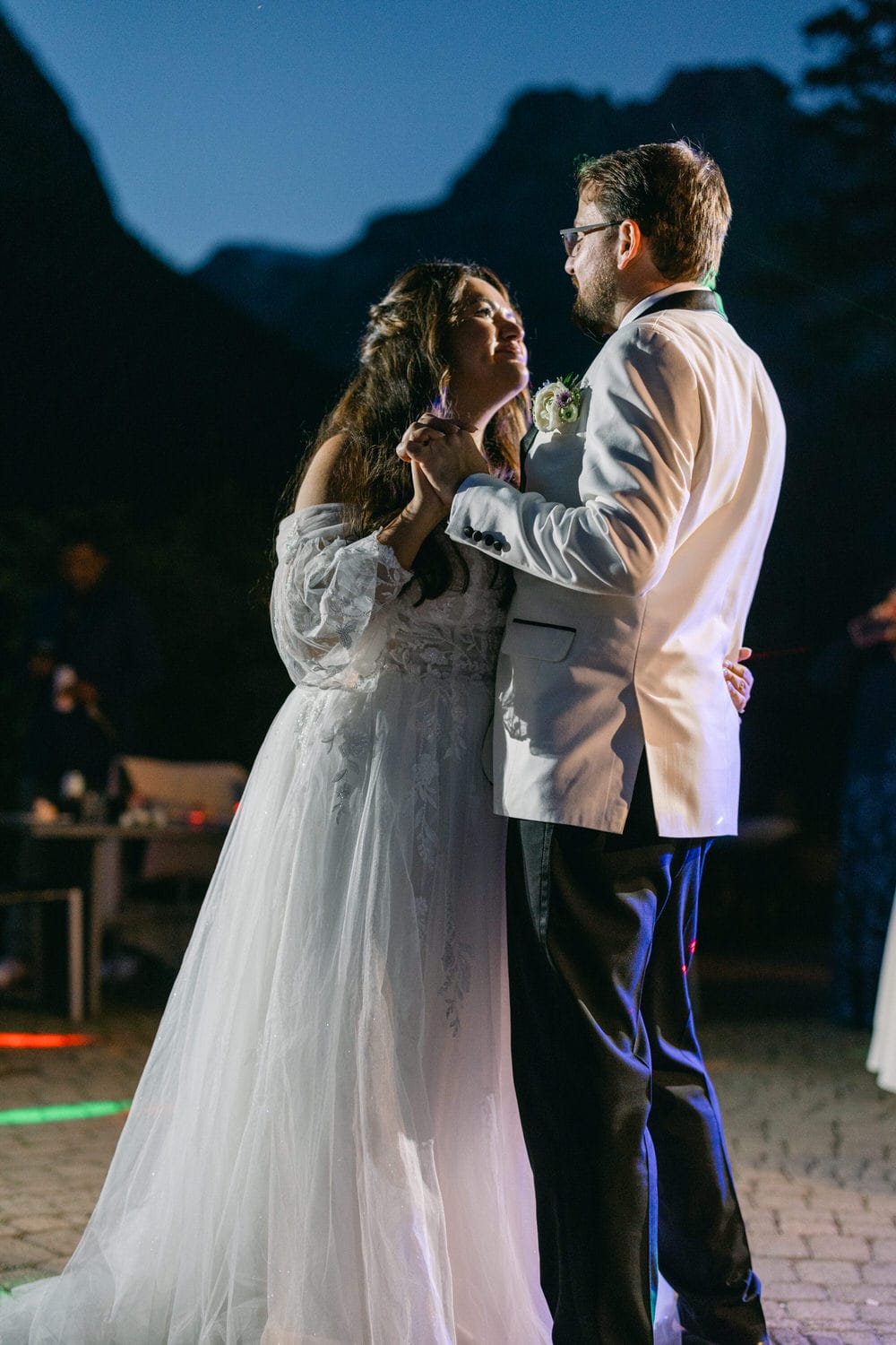 A bride and groom sharing a dance outdoors at twilight with mountains in the background.