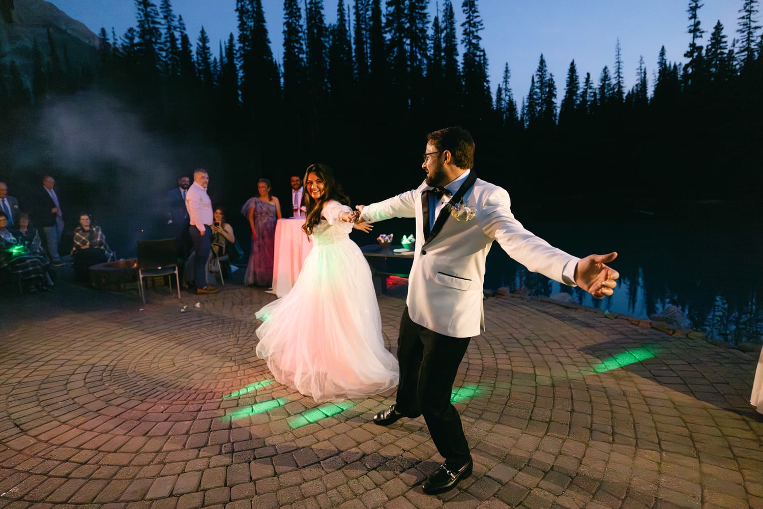 Bride and groom sharing a first dance outdoors with guests watching in a forested mountain setting at dusk.