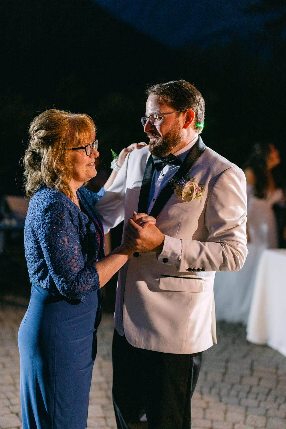 A woman in blue attire and a man in a cream-colored suit enjoying a dance together at an evening outdoor event.