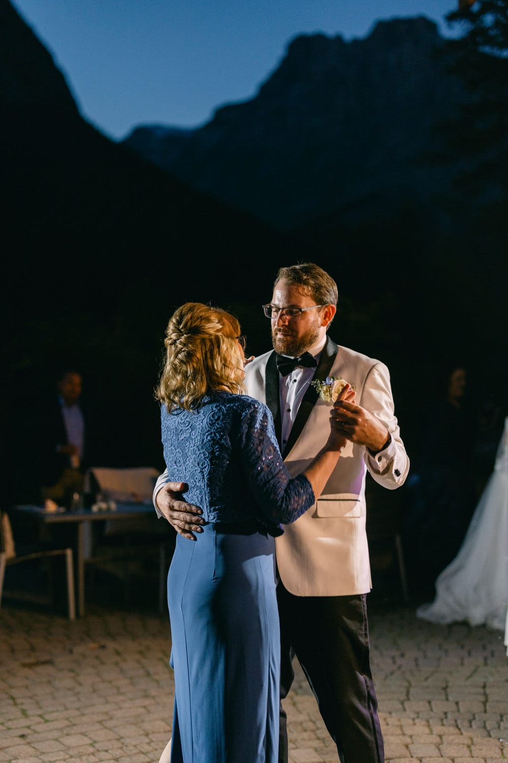 A man in a suit and a woman in a blue dress dancing together at an evening outdoor event with mountains in the background