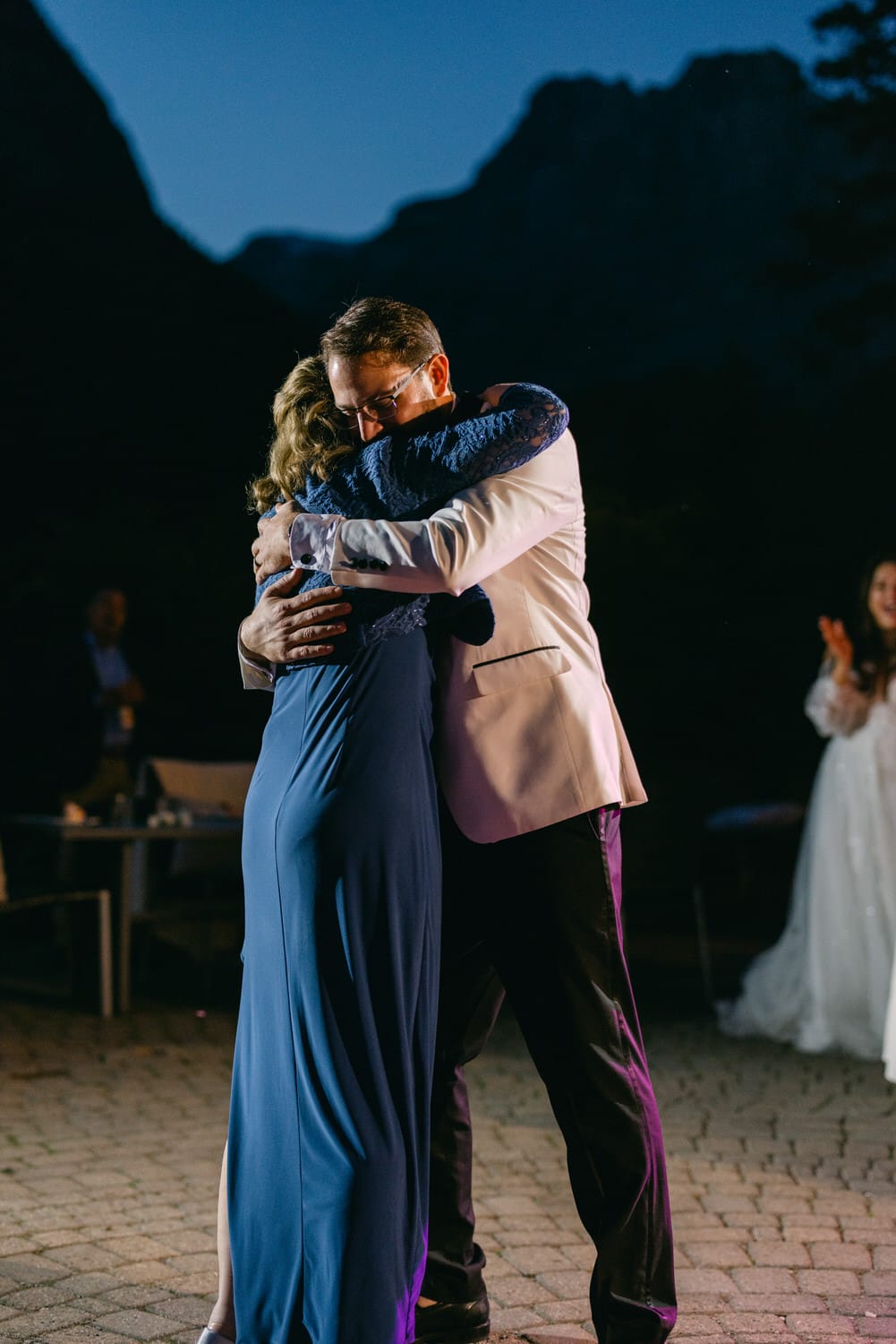 A man and woman sharing a heartfelt hug at an event with mountains in the background at dusk.