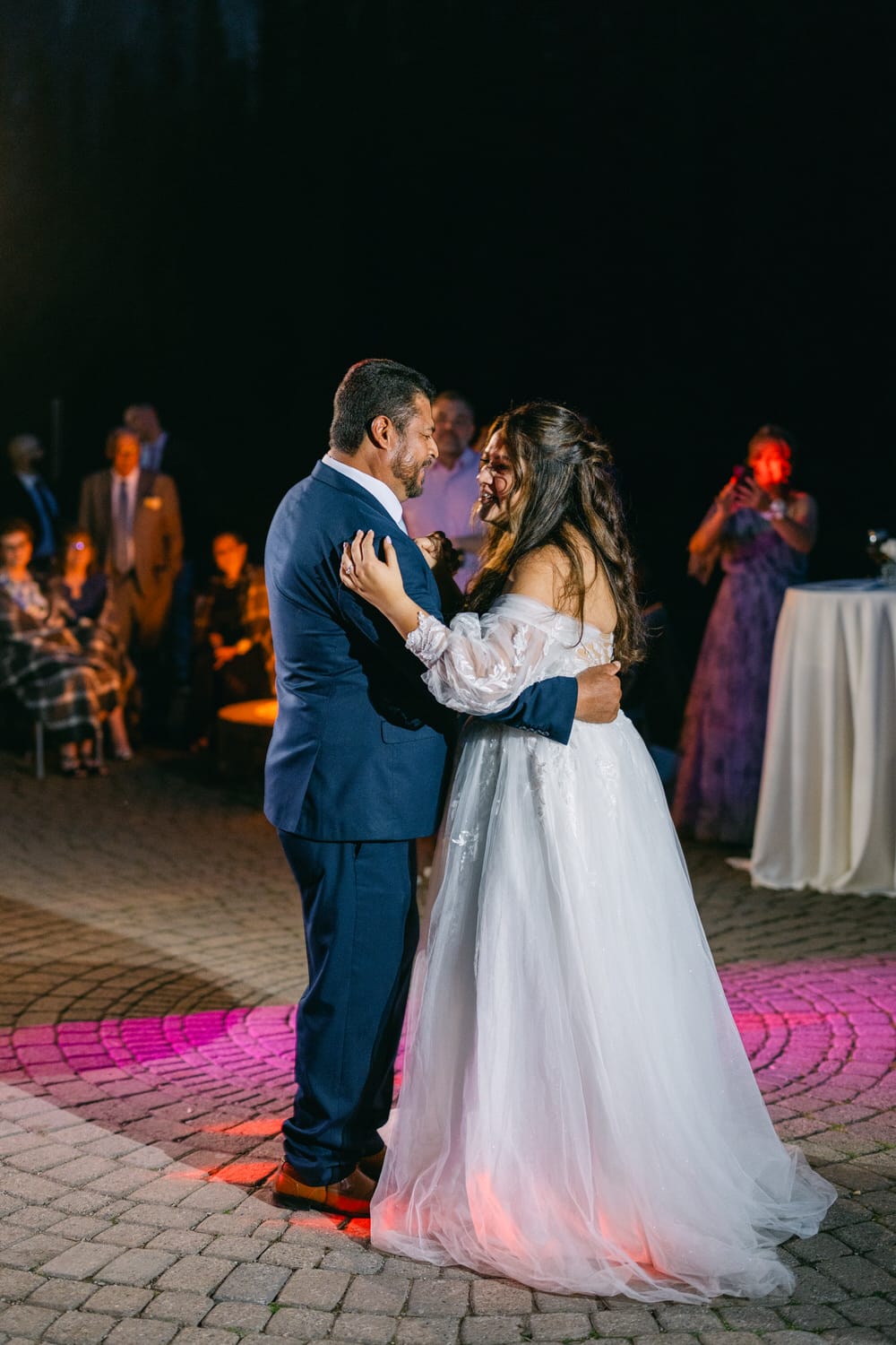 A bride and groom sharing a joyous dance outdoors at night, surrounded by guests.