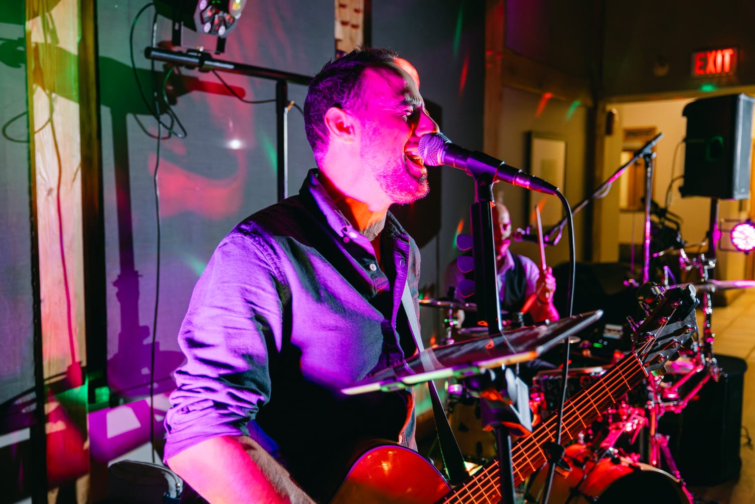 Guitarist singing into a microphone onstage with colorful stage lighting and drummer in the background.