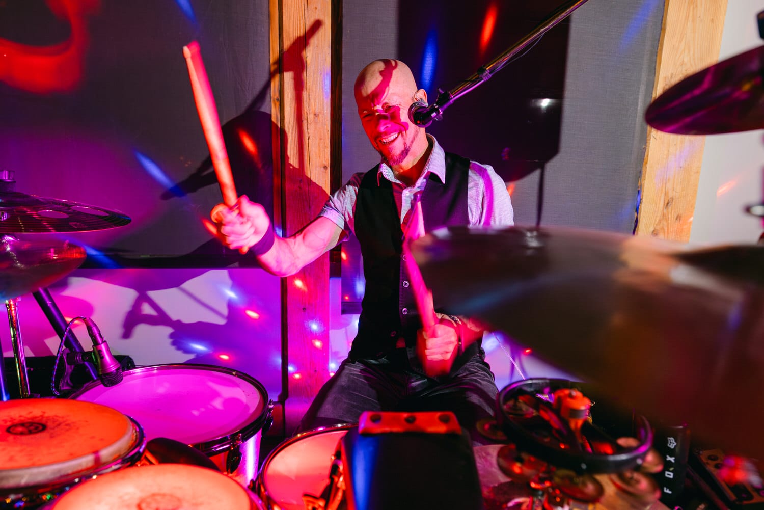 A bald musician in a vest and tie energetically playing the drums with colorful stage lights and shadows in the background.