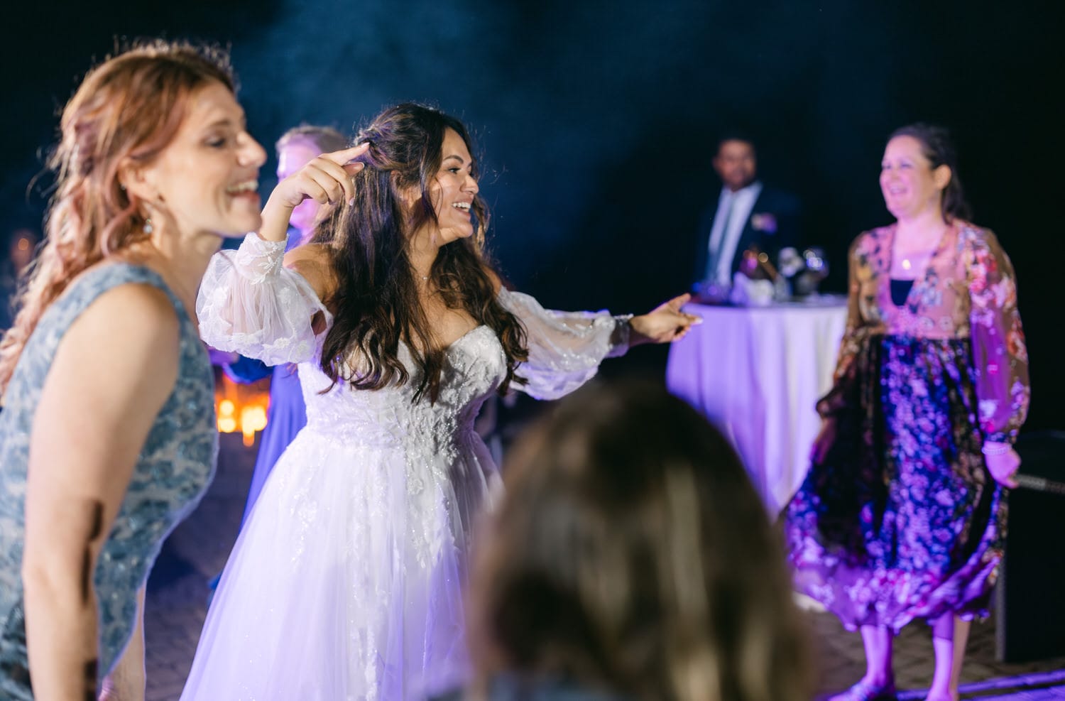 A bride happily dancing with guests at a wedding reception, candid moment captured with blurred lights in the background.