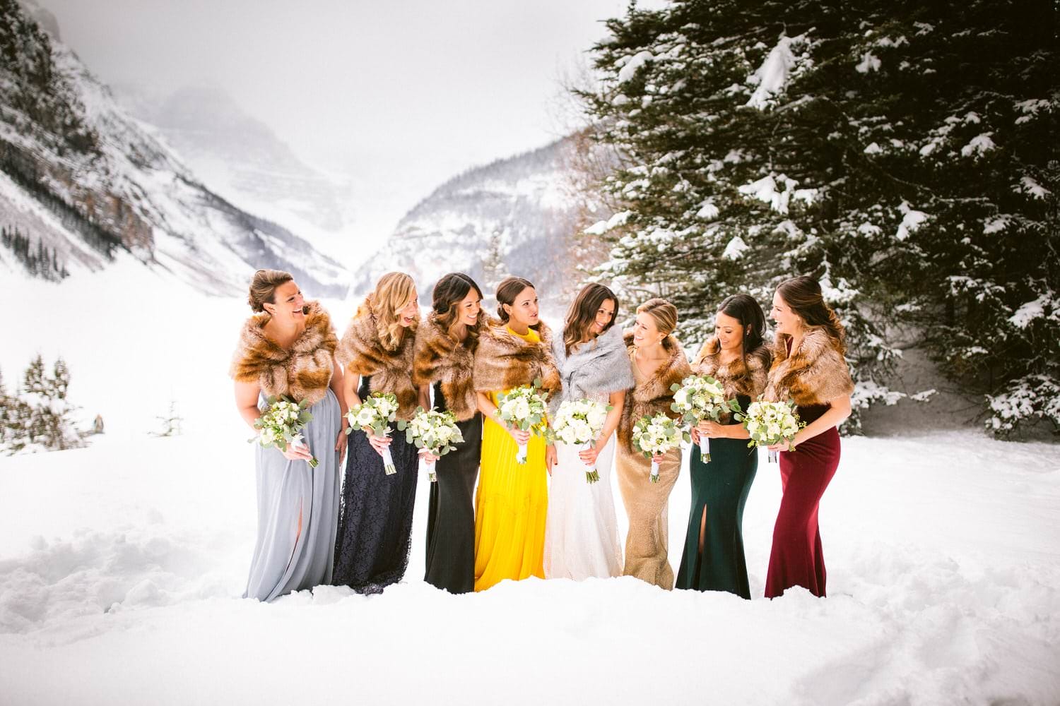 A group of bridesmaids in fur shawls and colorful dresses holding bouquets in a snowy mountain landscape