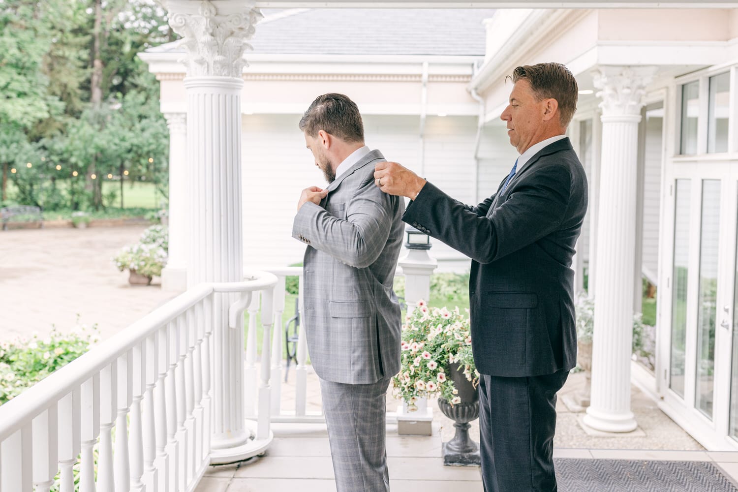 Two men in suits adjusting their jackets on a porch with a garden backdrop.