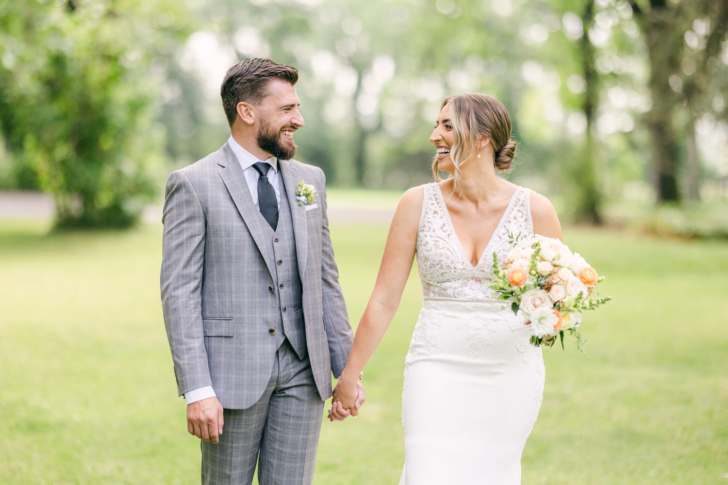 A smiling bride and groom holding hands and looking at each other in a green park setting, with the bride holding a bouquet of flowers.