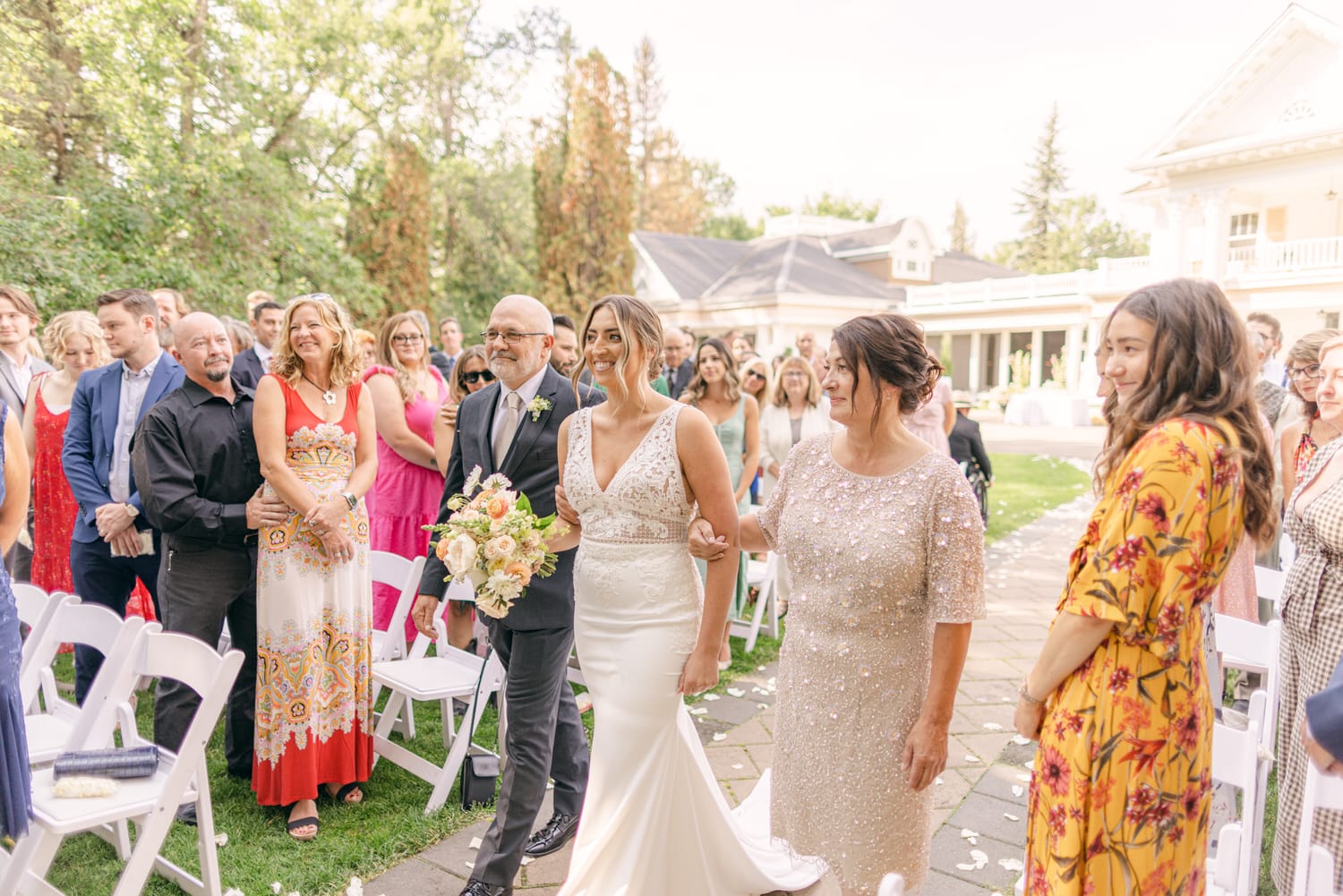 A smiling bride walking down the aisle with her parents, guests standing and watching during an outdoor wedding ceremony.