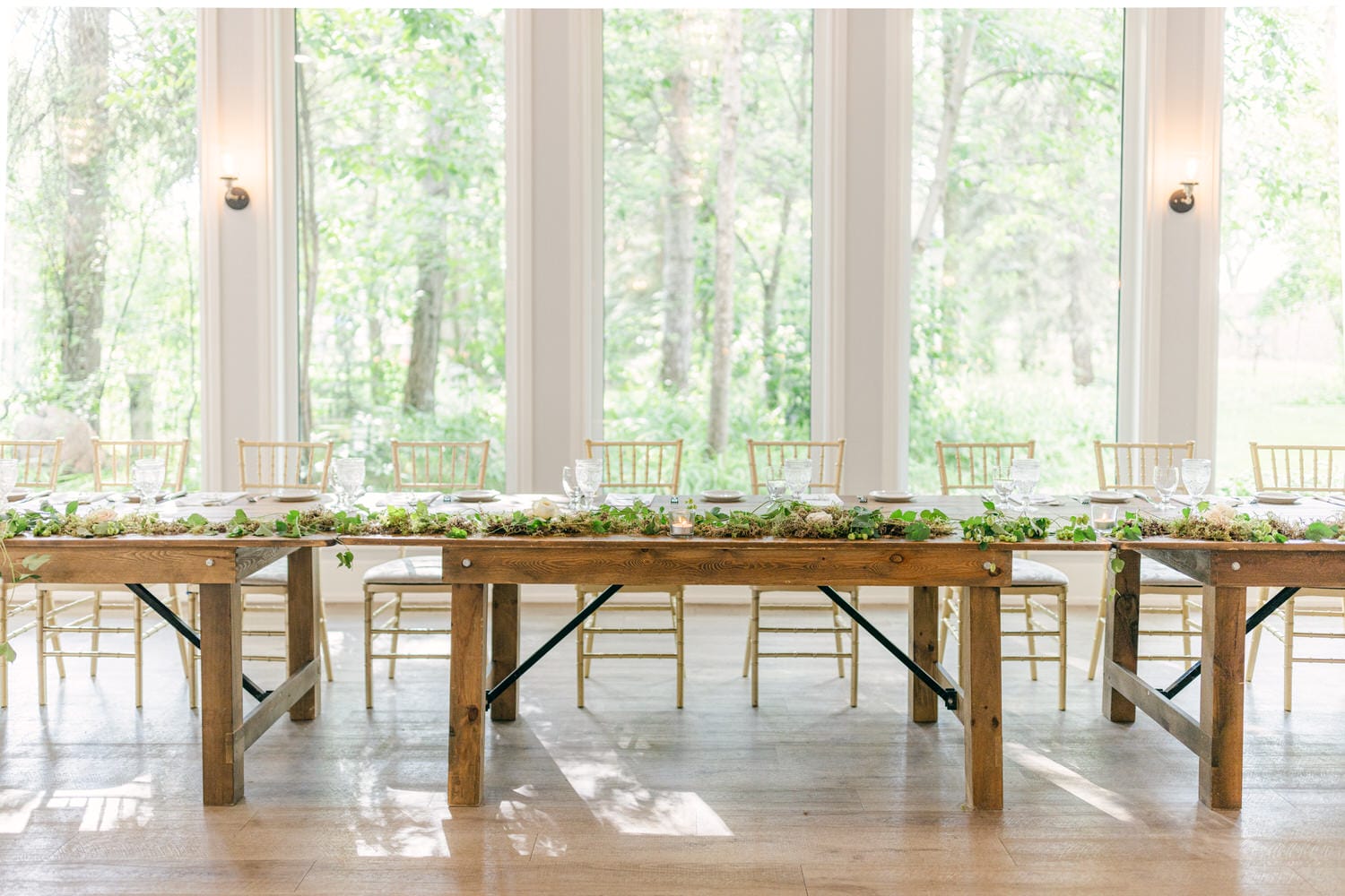 A bright and airy wedding reception hall with wooden tables, elegant place settings, and lush greenery centerpiece decorations on a sunny day.