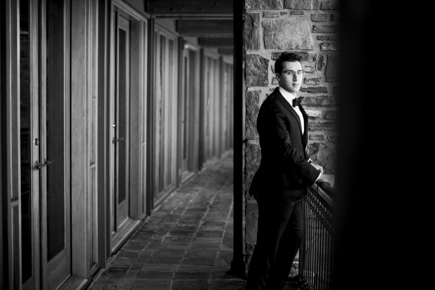 A black and white photo of a man in a suit standing by a railing in a corridor with doors in the background.