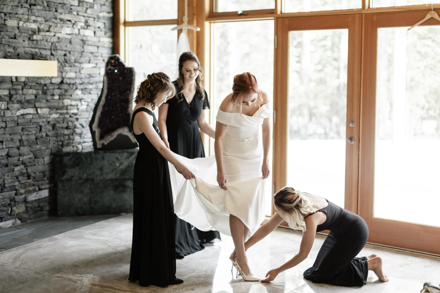 A bride in a white dress being helped by three women, with one adjusting her shoe, in a room with large windows and a stone wall.