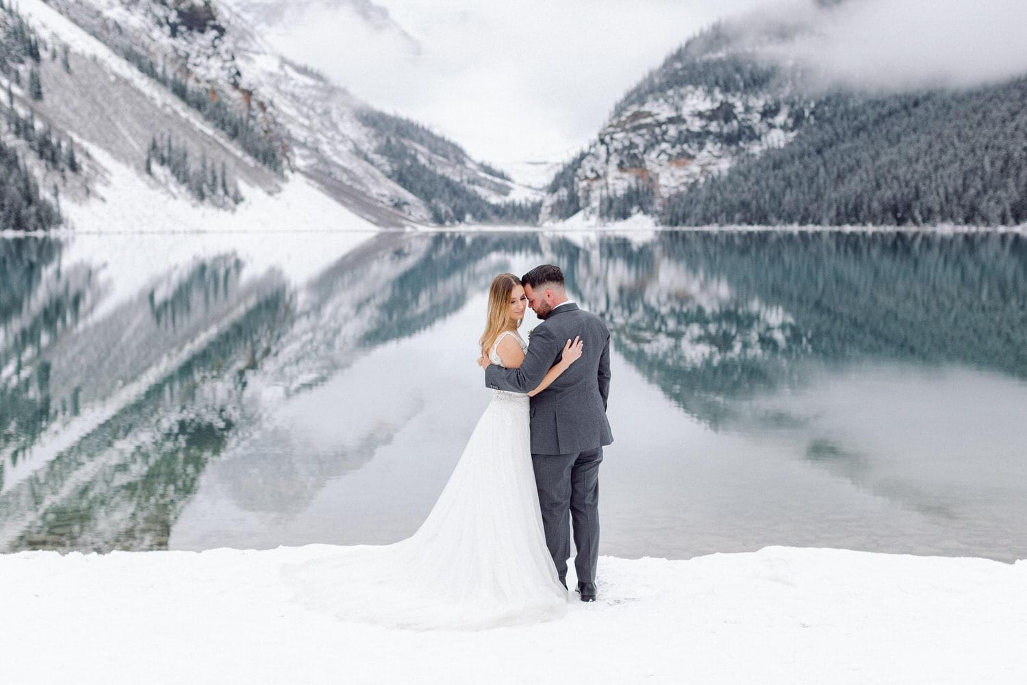 A bride and groom embracing on the snowy shore of a lake with mountains and reflections in the background.