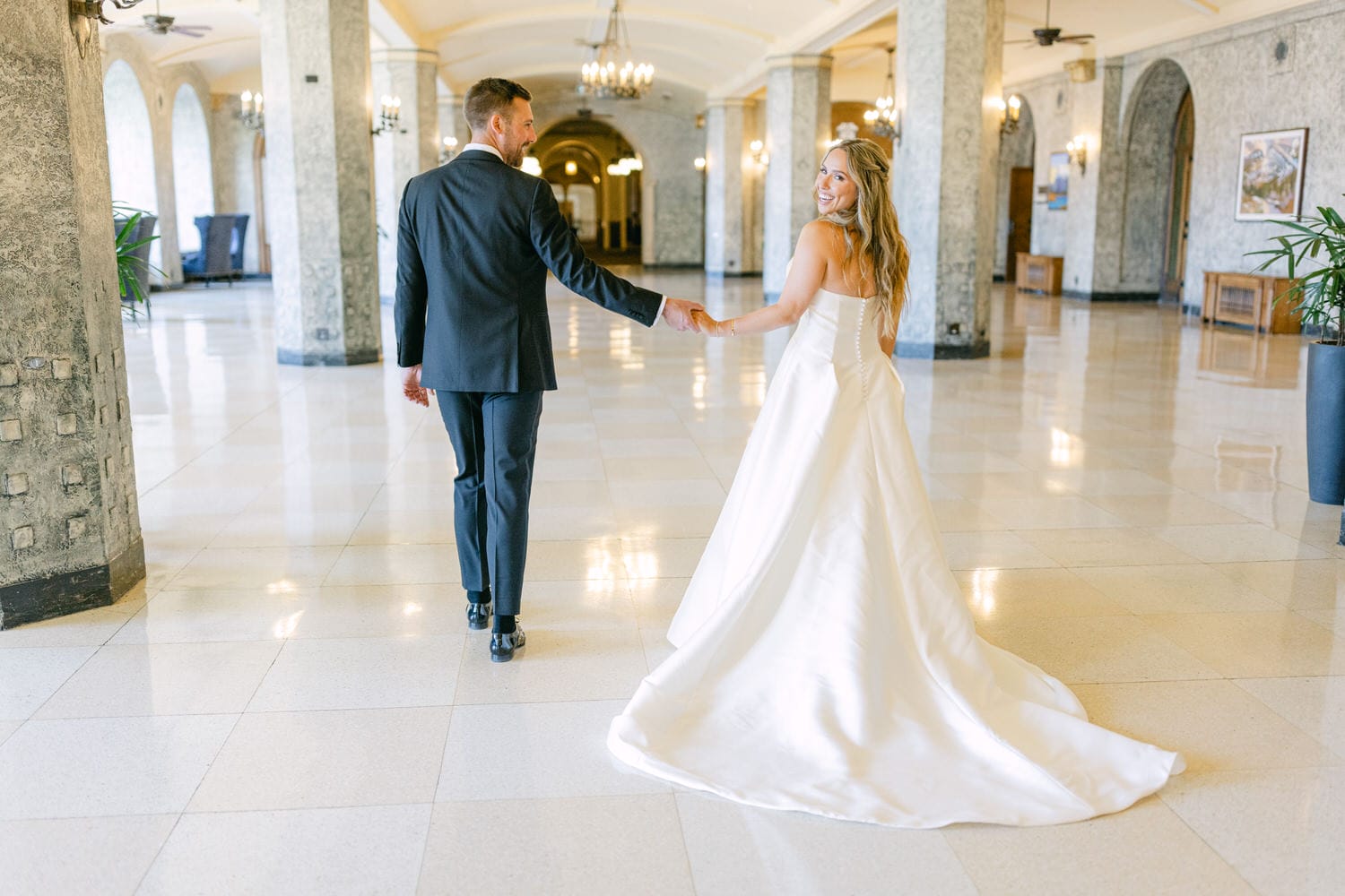 A bride in a white dress and a groom in a suit holding hands and looking at each other with smiles in a grand hallway.