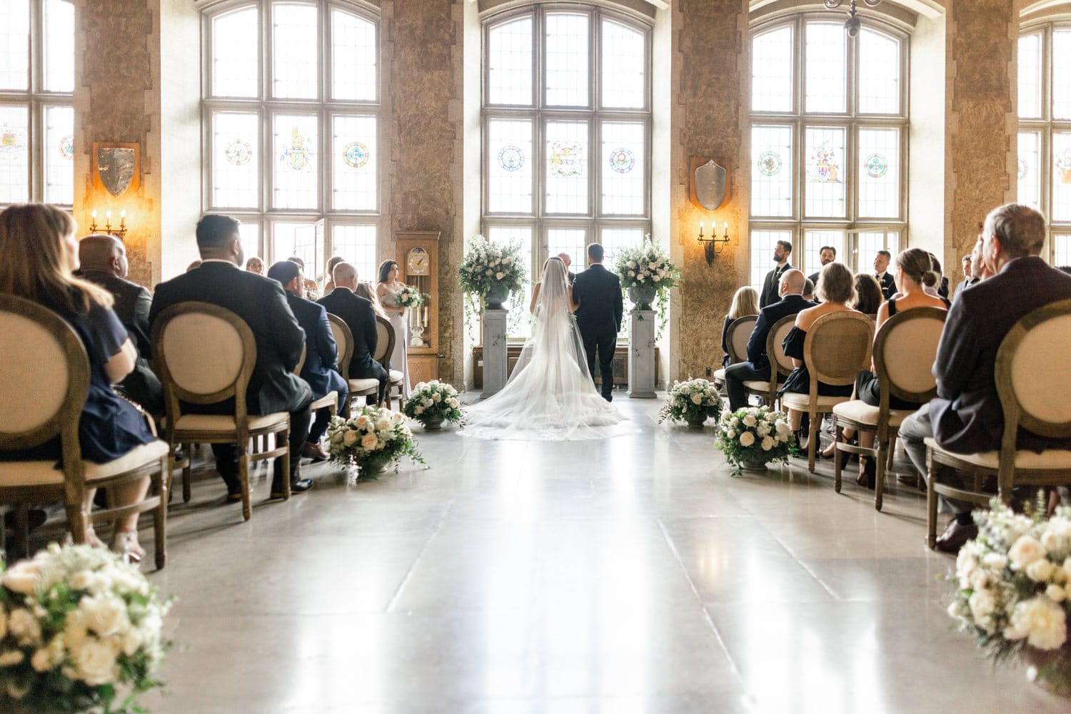 A bride and groom standing at the altar during a wedding ceremony, with guests seated on either side and sunlight streaming through large windows.