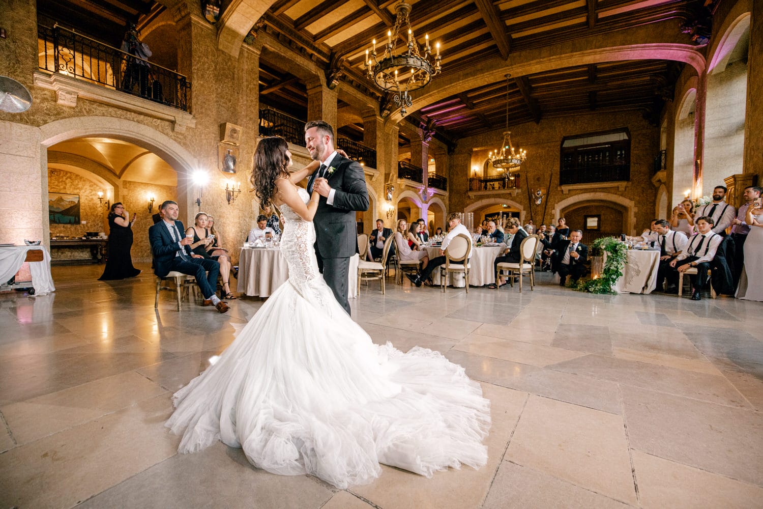 Bride and groom sharing their first dance in a grand ballroom with guests looking on.