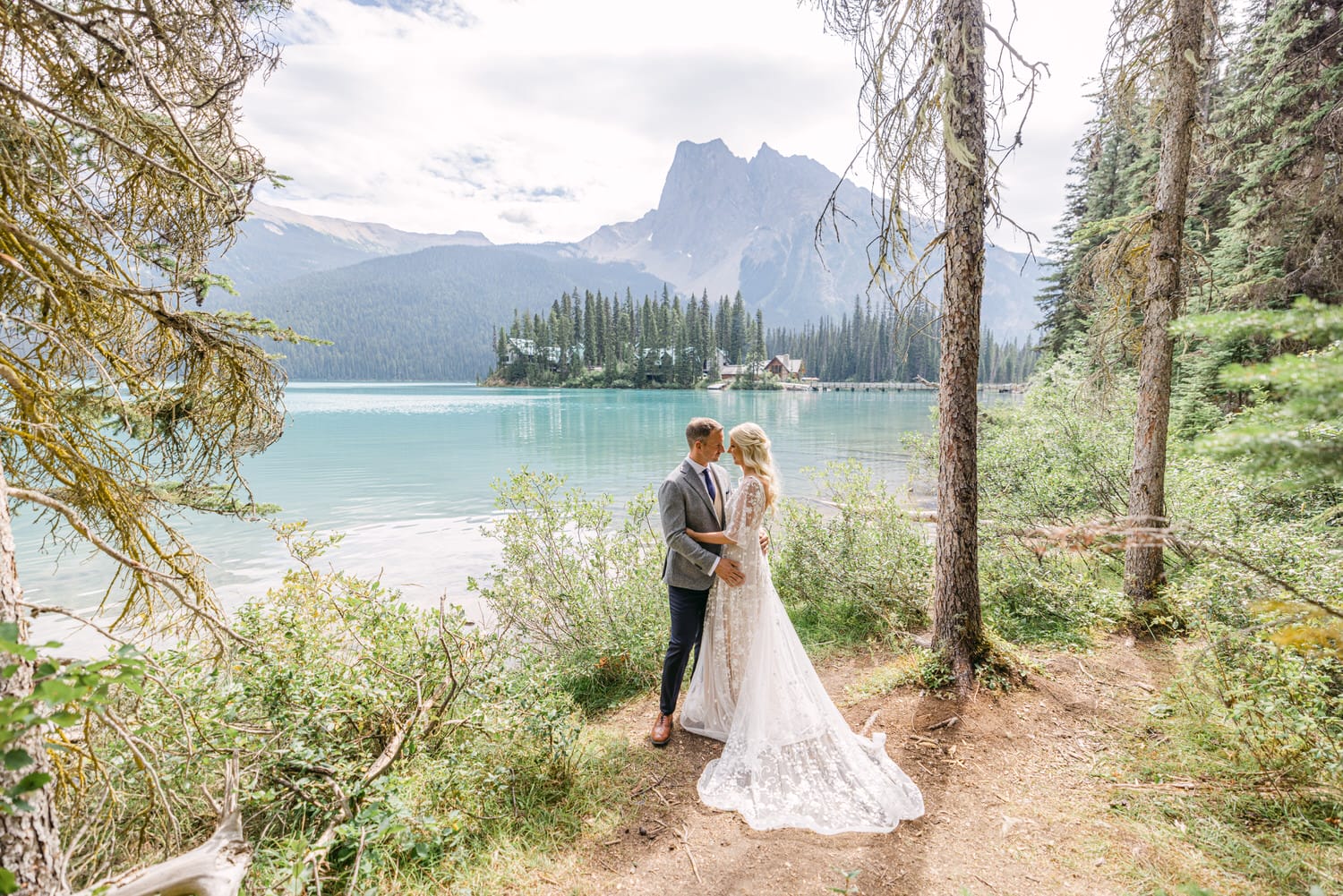 A couple in wedding attire embracing by a serene mountain lake surrounded by forest.