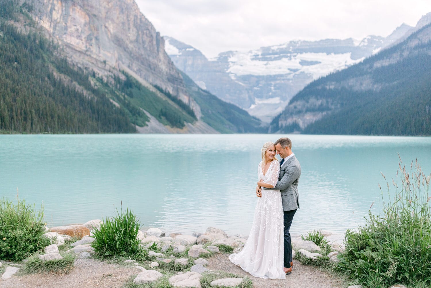 A couple in wedding attire sharing a moment by a serene mountain lake with majestic mountain cliffs and a glacier in the background.