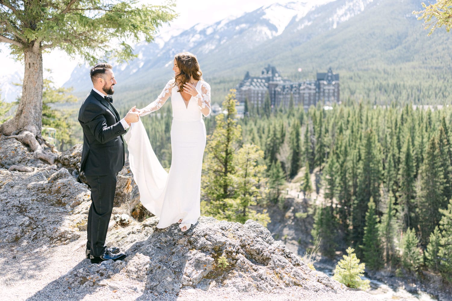 A bride and groom holding hands on a rocky outcrop with a panoramic view of mountains and forest in the background.