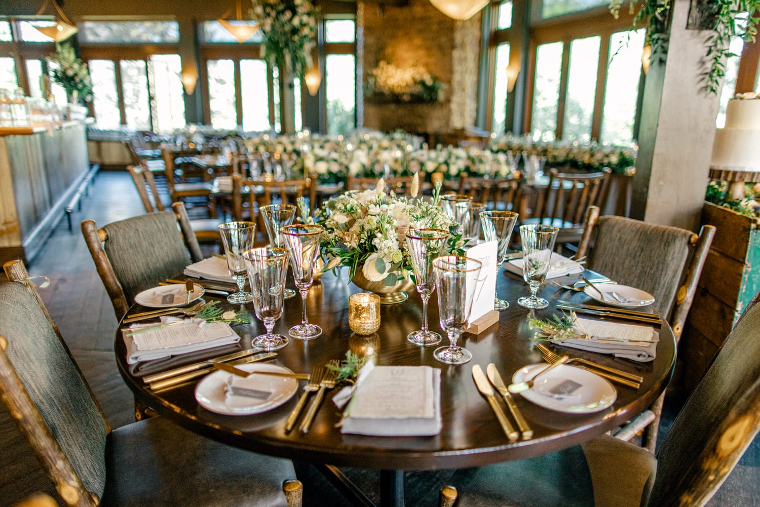 A beautifully arranged wedding reception table with floral centerpiece, glassware, and place settings in a venue with large windows and ambient lighting.