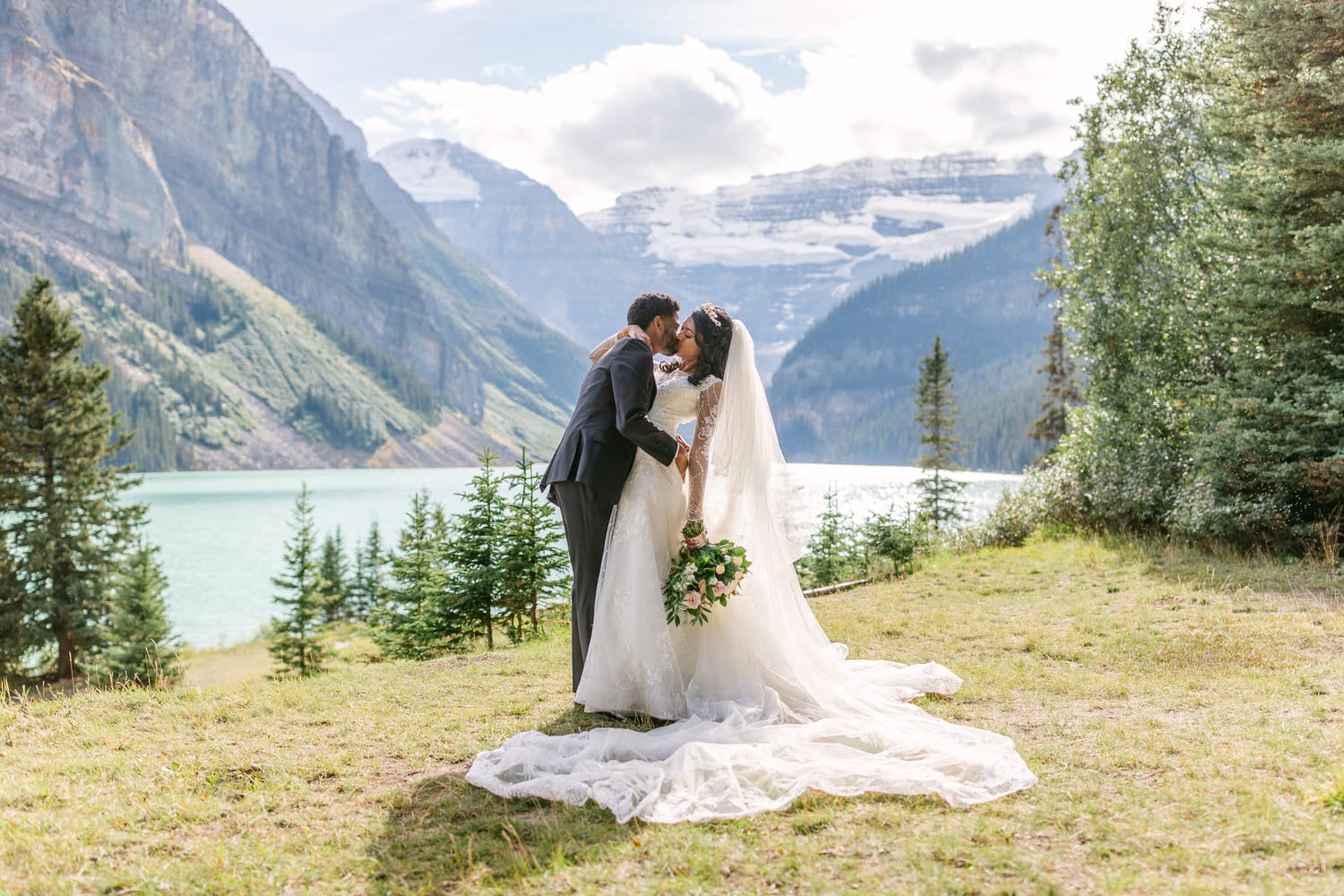 A bride and groom share a kiss in a picturesque mountain landscape by a lake, with the bride holding a bouquet and wearing a long flowing veil.