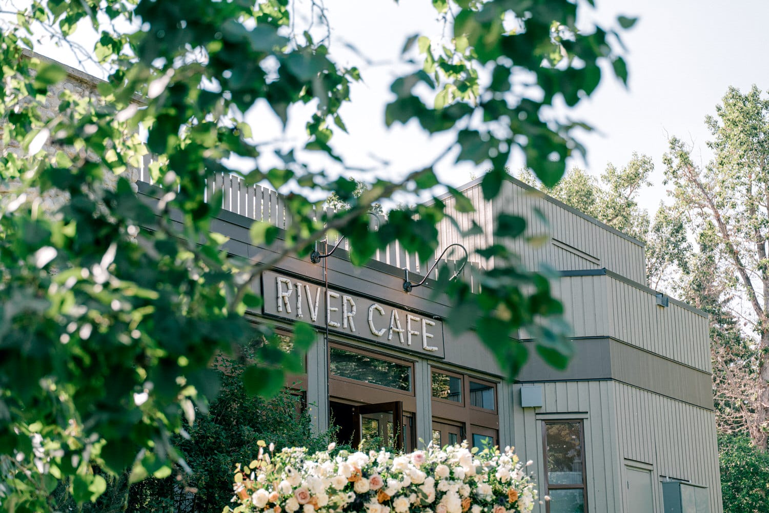 Exterior view of River Cafe surrounded by green foliage with a clear blue sky in the background.