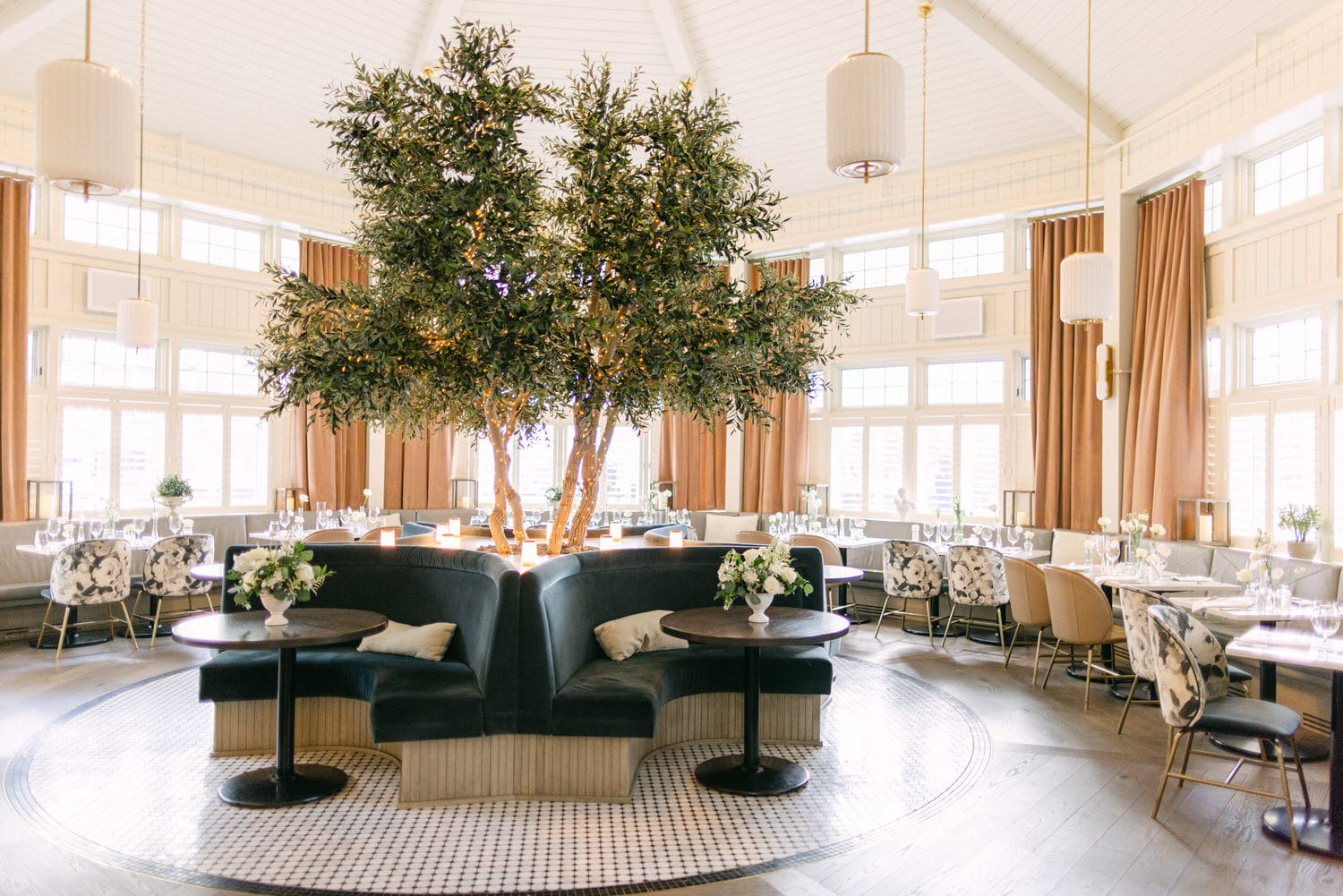 An expansive room adorned with a large indoor tree illuminated with fairy lights, surrounded by stylish furnishings and dining tables set with white flowers.