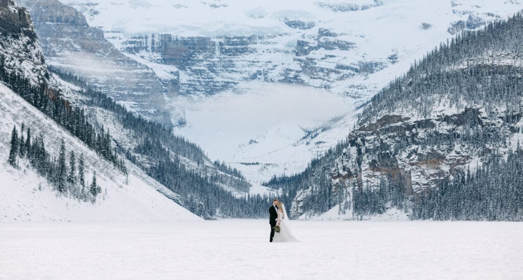 A couple embracing in the center of a snowy landscape with towering mountains in the background.