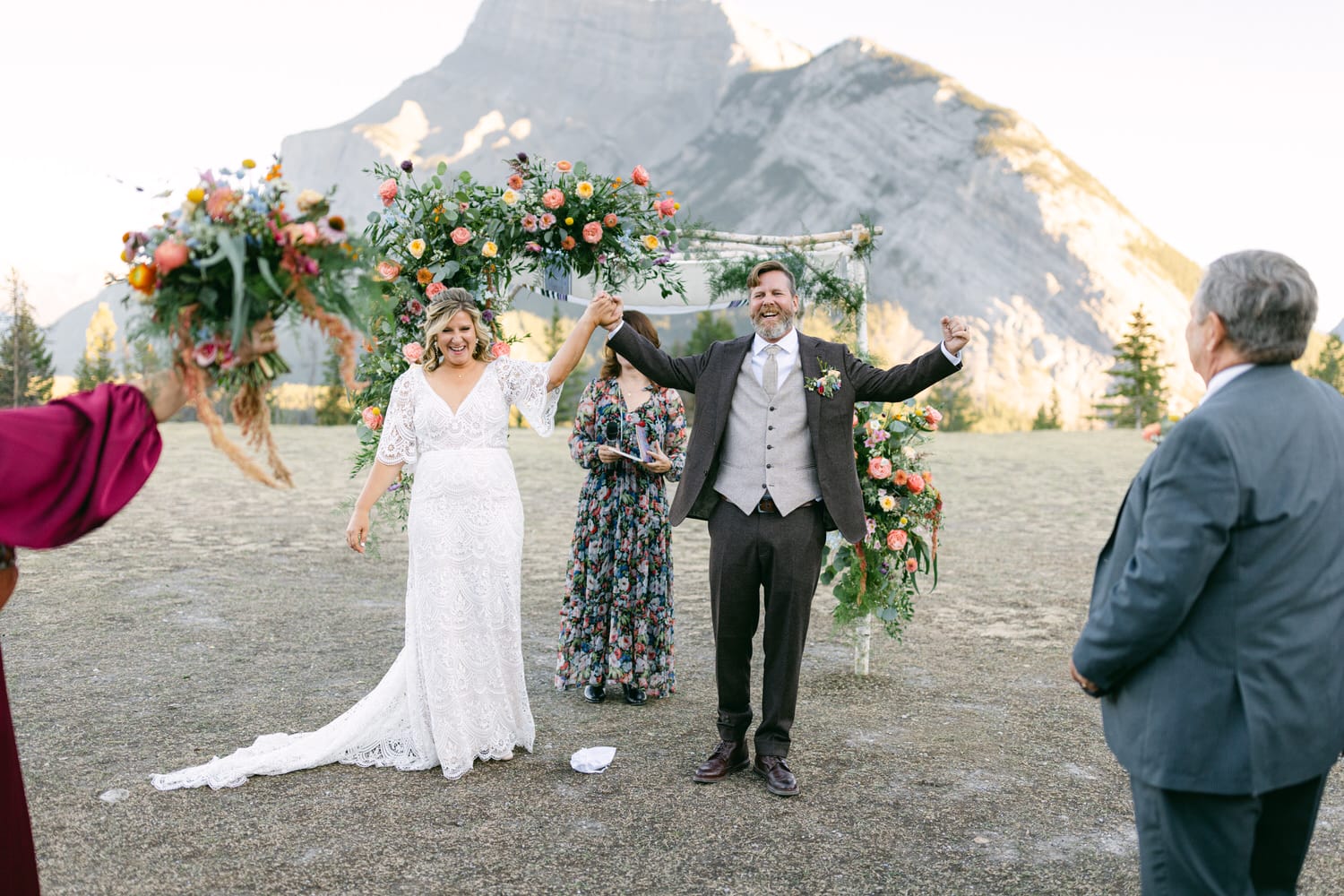 A bride and groom happily exiting their wedding ceremony with their hands raised under a floral arch, with mountains in the background.