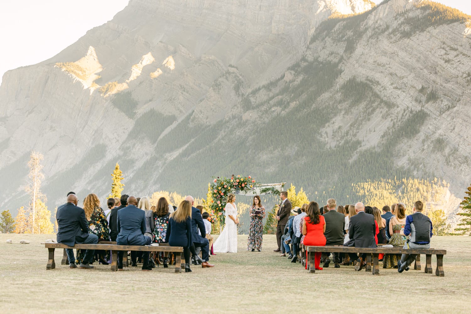 Guests seated on benches at an outdoor wedding ceremony with a mountainous backdrop, featuring a couple standing under a floral arch.