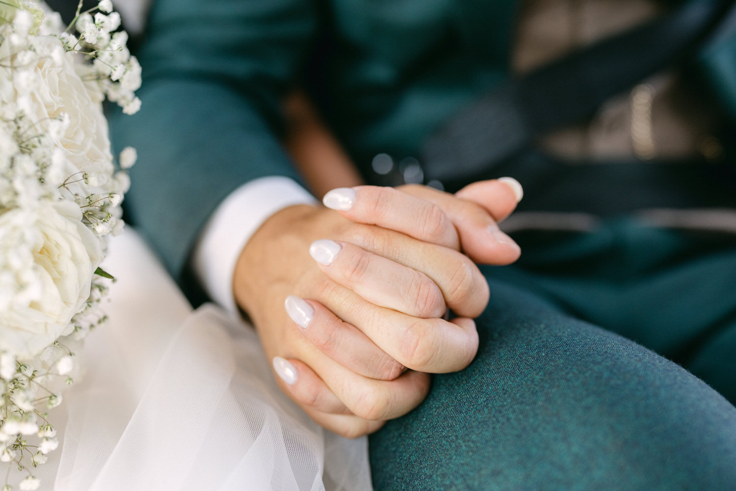 A close-up of two hands clasped together, one adorned with polished nails, alongside a wedding bouquet featuring white flowers and baby's breath, set against the backdrop of formal attire.