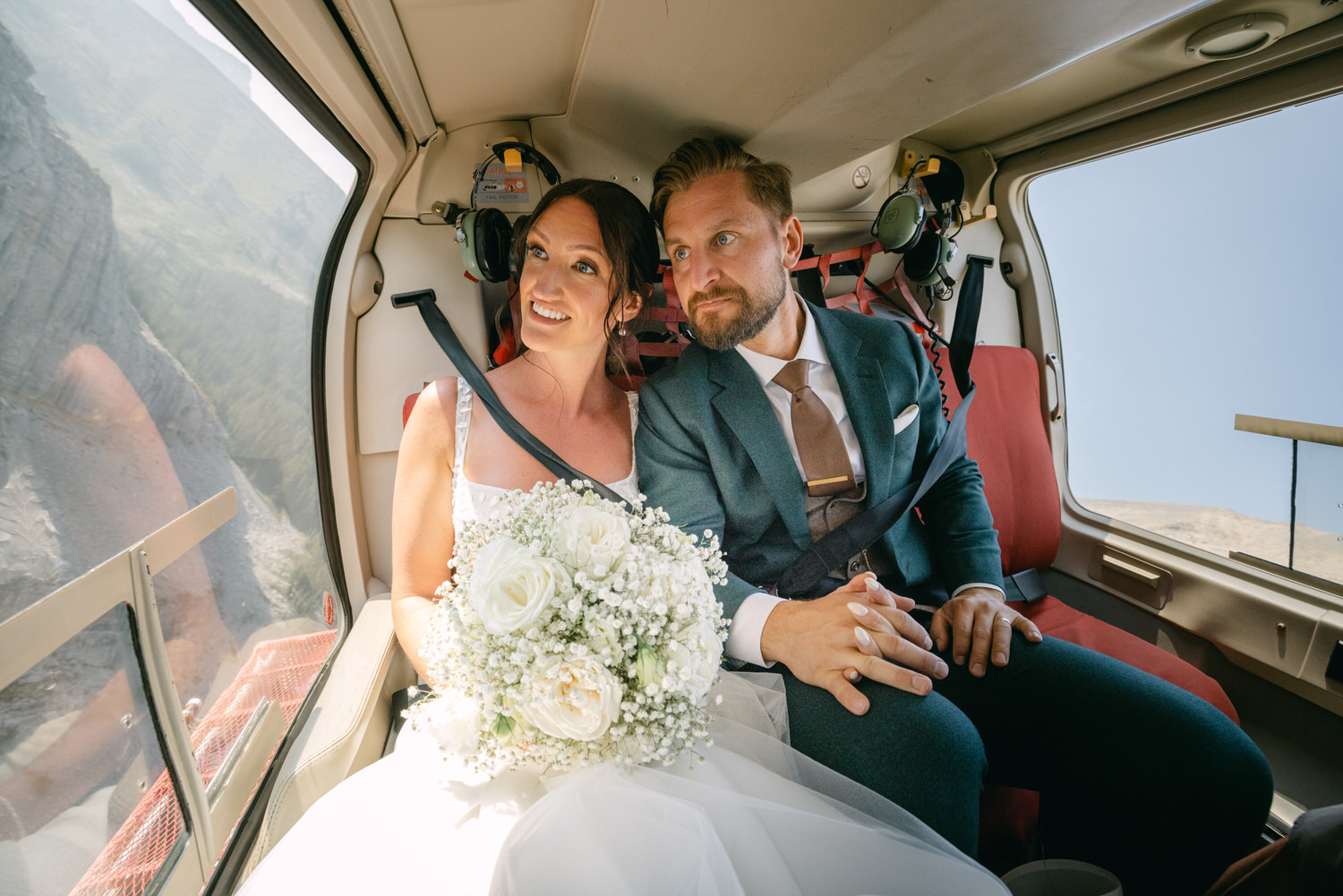 A bride and groom sharing a joyful moment inside a helicopter, with the bride holding a bouquet and the groom looking at her affectionately while enjoying a scenic view through the window.