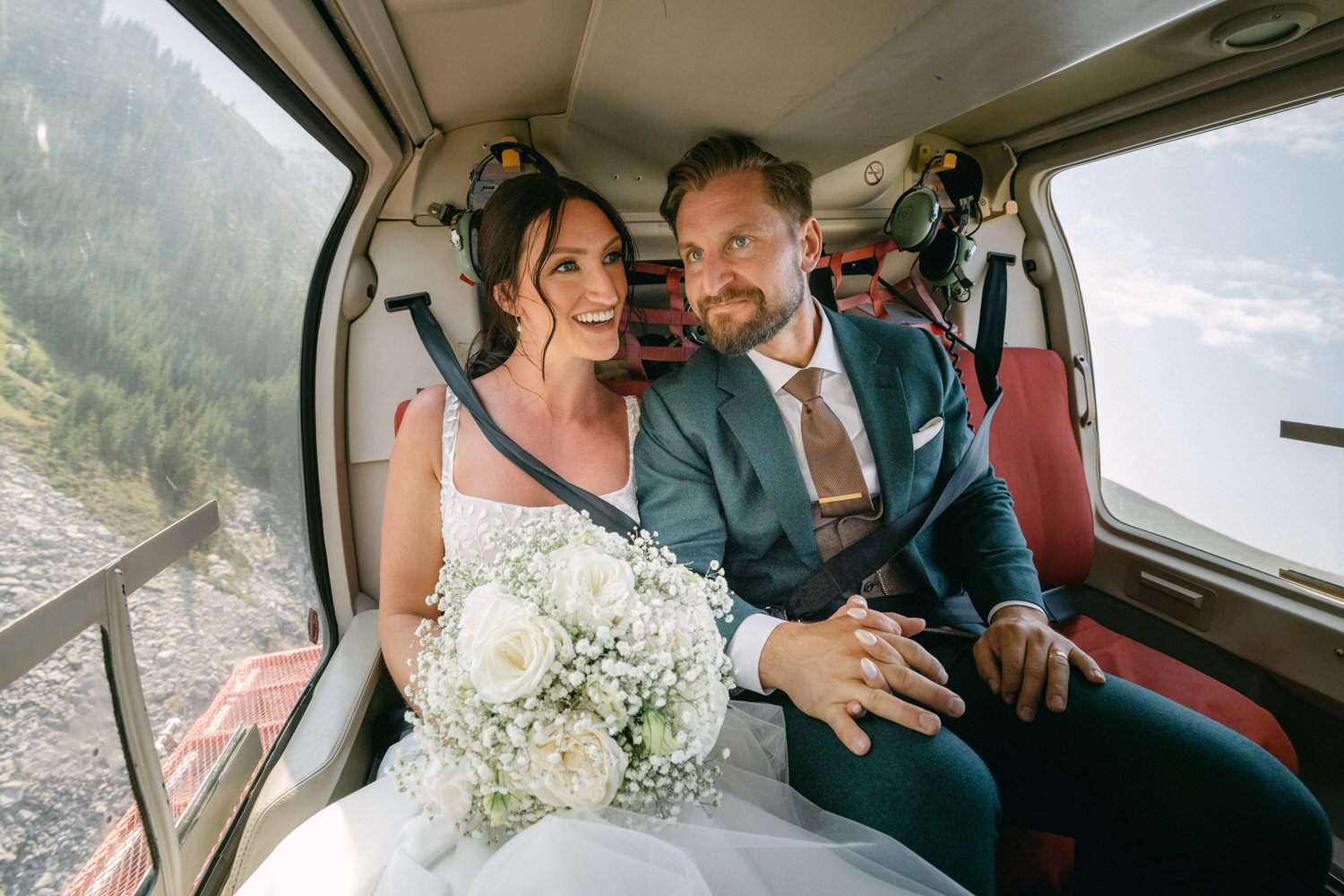 A joyful bride and groom sit closely in a helicopter, surrounded by stunning natural scenery, as they share a loving moment and hold hands while the bride holds a bouquet of white roses.