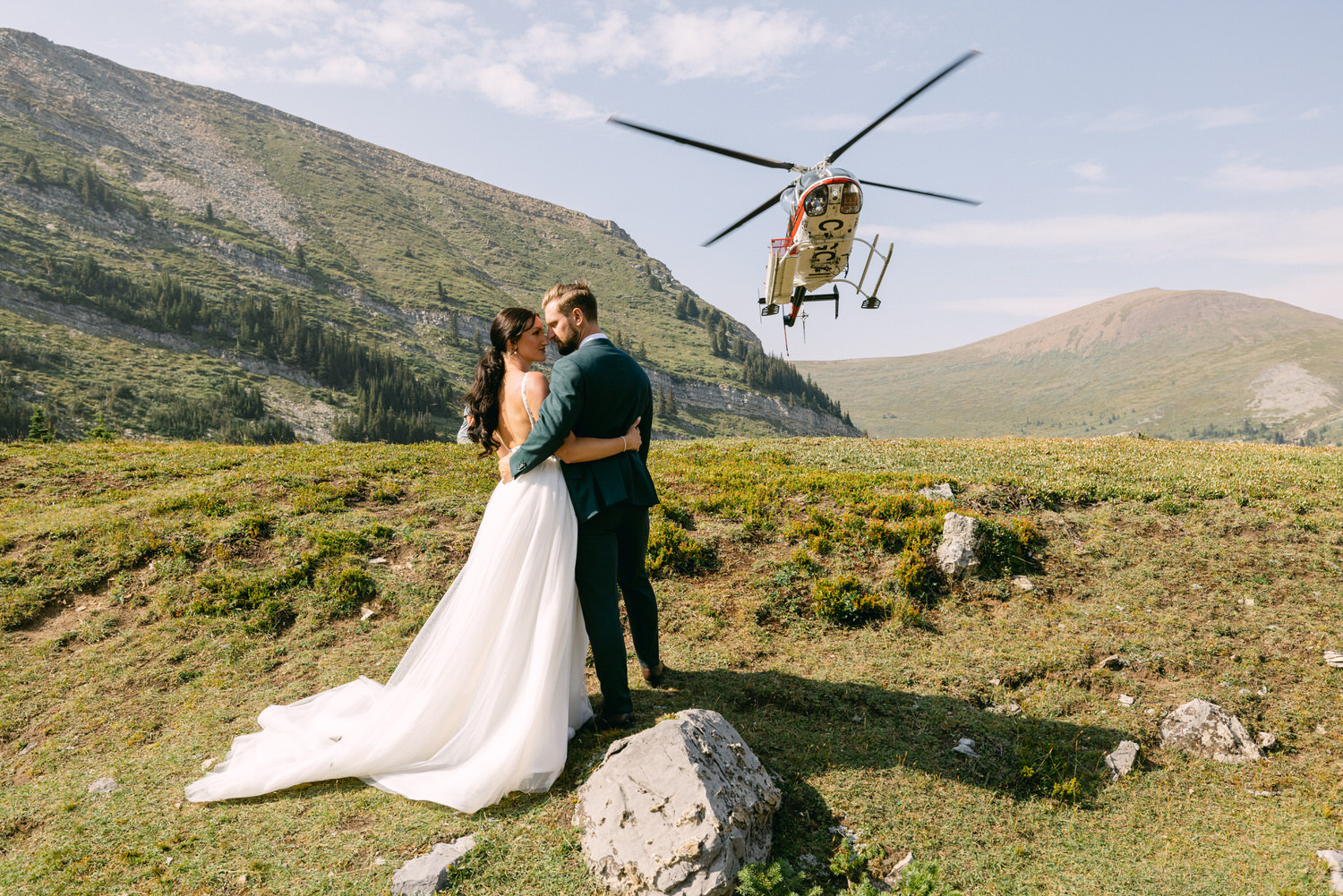 A couple embraces in a picturesque mountain setting as a helicopter flies above them, showcasing their unique wedding backdrop.