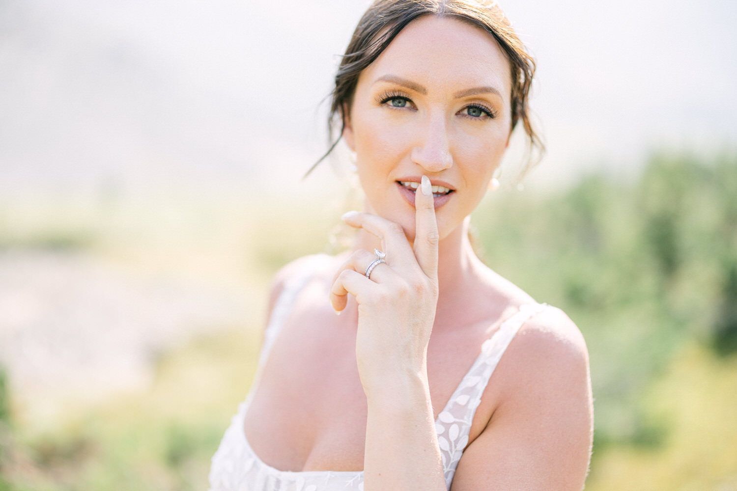 A close-up of a woman in a wedding dress, playfully holding a finger to her lips outdoors, with a soft focus on a natural background.