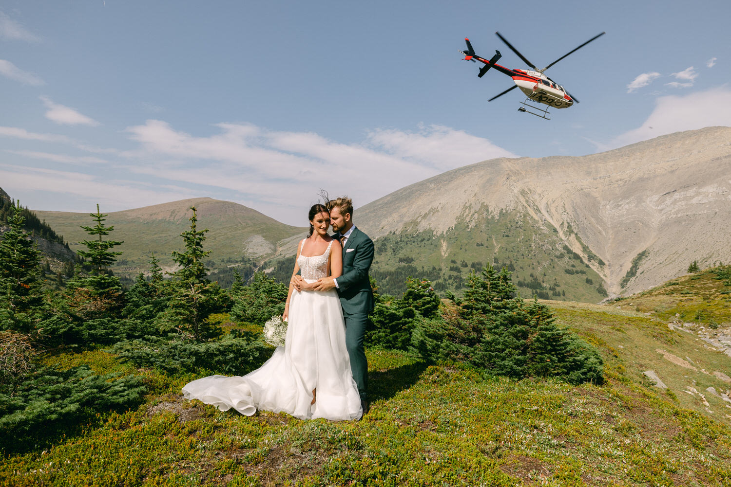 A couple in wedding attire embraces in a scenic mountain landscape, with a helicopter flying overhead amidst greenery and rocky hills.