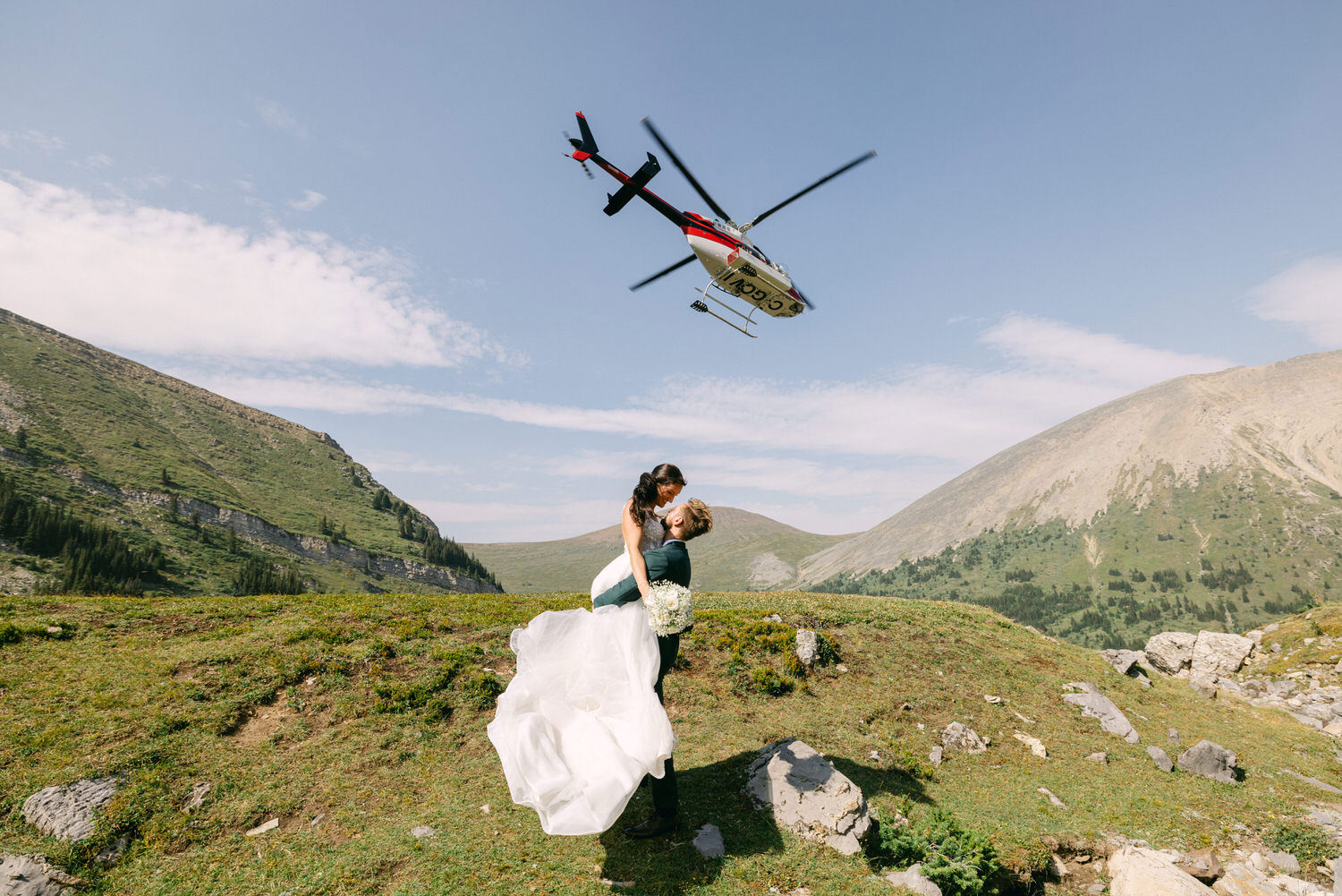 A couple shares a romantic kiss with a helicopter flying overhead in a stunning mountainous landscape.