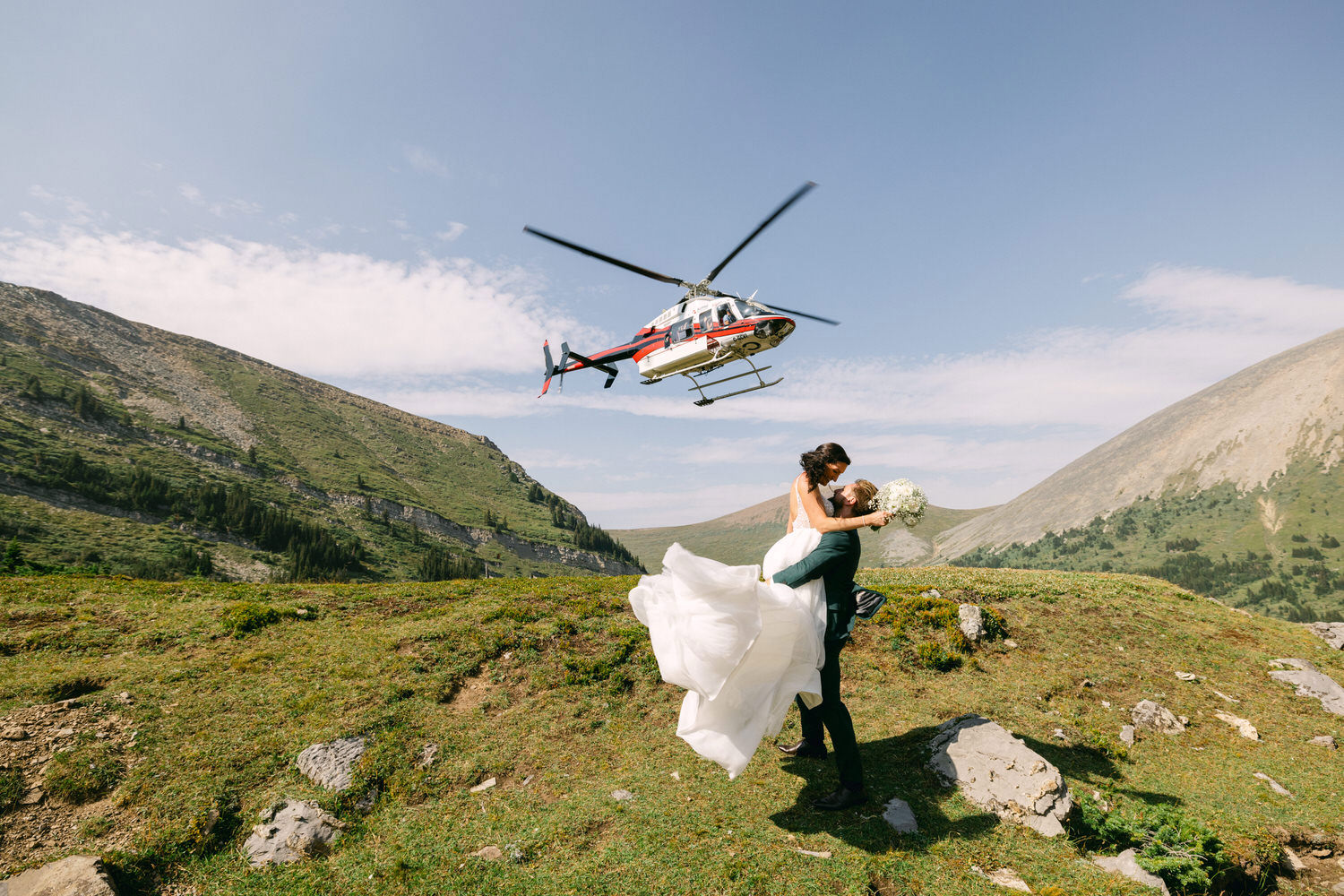 A newlywed couple embraces in a mountainous landscape as a helicopter flies overhead, capturing a moment of joy on their special day.