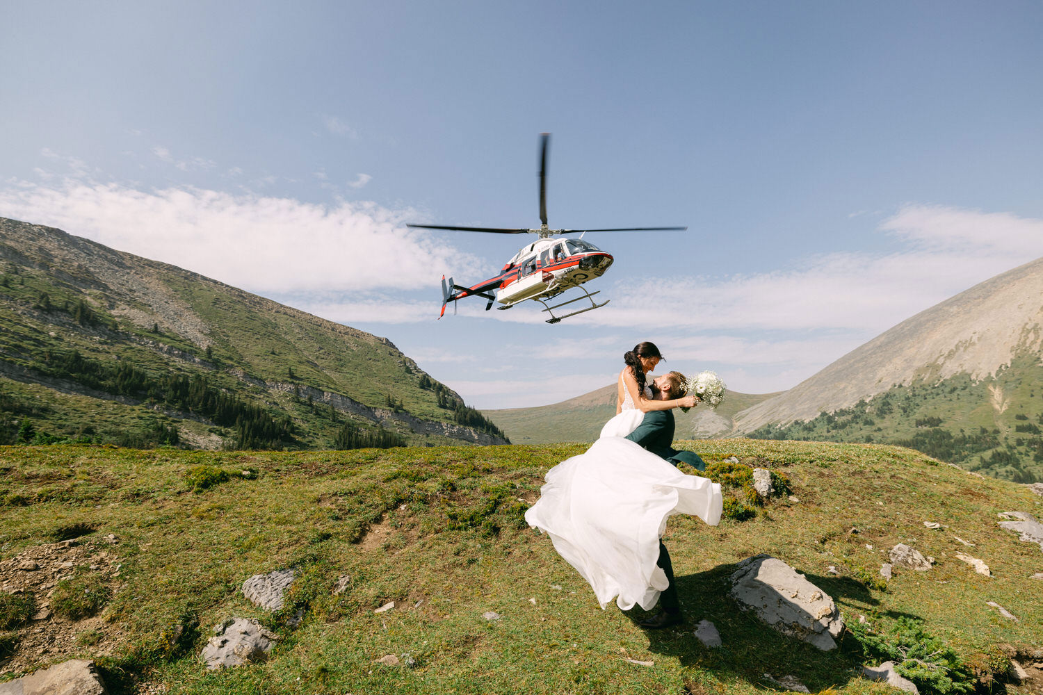 A bride and groom celebrate their love as a helicopter hovers above them in a scenic mountain landscape.